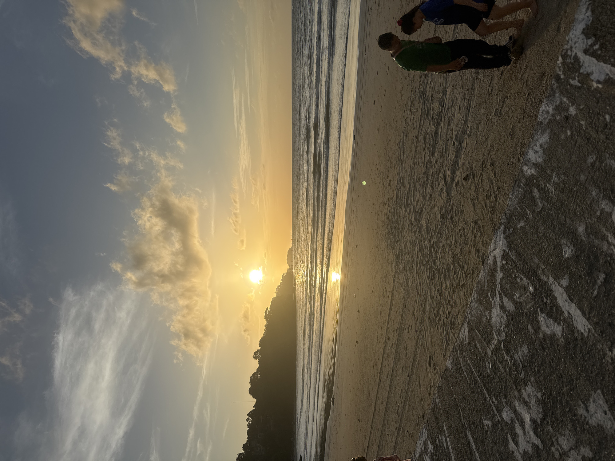 The image depicts a sunset scene on the beach in Patong, Thailand. The sun is low on the horizon, casting a golden glow over the ocean and the sand. The sky is a gradient of orange and blue, with scattered clouds. Gentle waves are rolling onto the shore.
In the foreground, a concrete pathway or structure runs along the edge of the sand. On the sand, a man wearing a green t-shirt and dark pants stands next to a young woman in a blue t-shirt and dark shorts. The man holds a clear bottle of water. They appear to be looking down or conversing. Further to the left, a portion of another person's leg, wearing a pink garment, is visible at the edge of the frame.
The background features a treed hillside rising from the beach, with buildings and a tall structure visible near the summit. A pier or jetty extends into the water in the distance on the right. The overall impression is of a peaceful evening on a tropical beach.