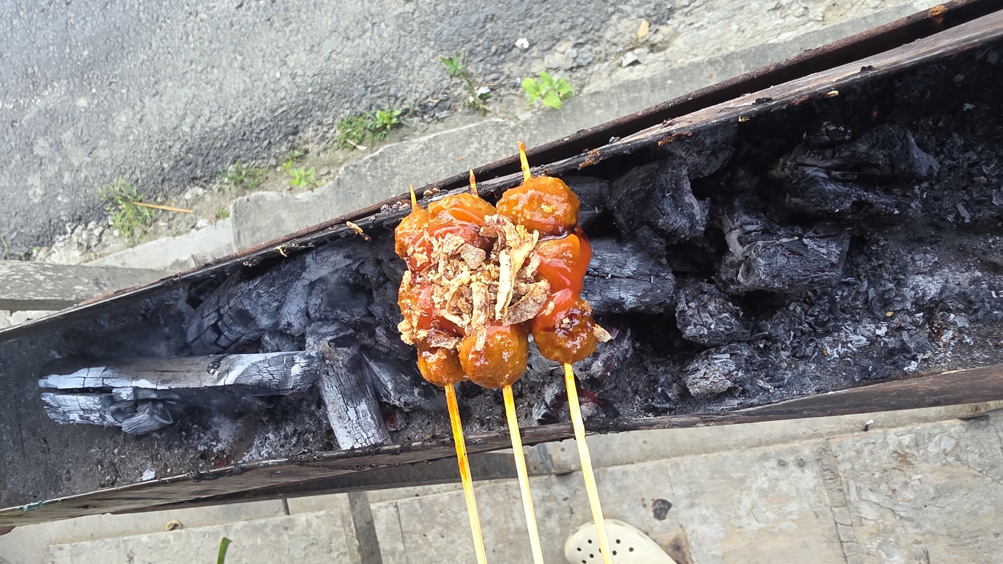 The image shows three skewers of meatballs, covered in sauce and fried onions, resting on a charcoal grill. The grill is made of rusty metal and filled with burnt charcoal and wood. The location appears to be outdoors, possibly near a street, as asphalt is visible in the background. In the lower right corner, a white shoe is partially visible. Given the prompt, the scene is potentially located in the city of Lhokseumawe, Indonesia.
