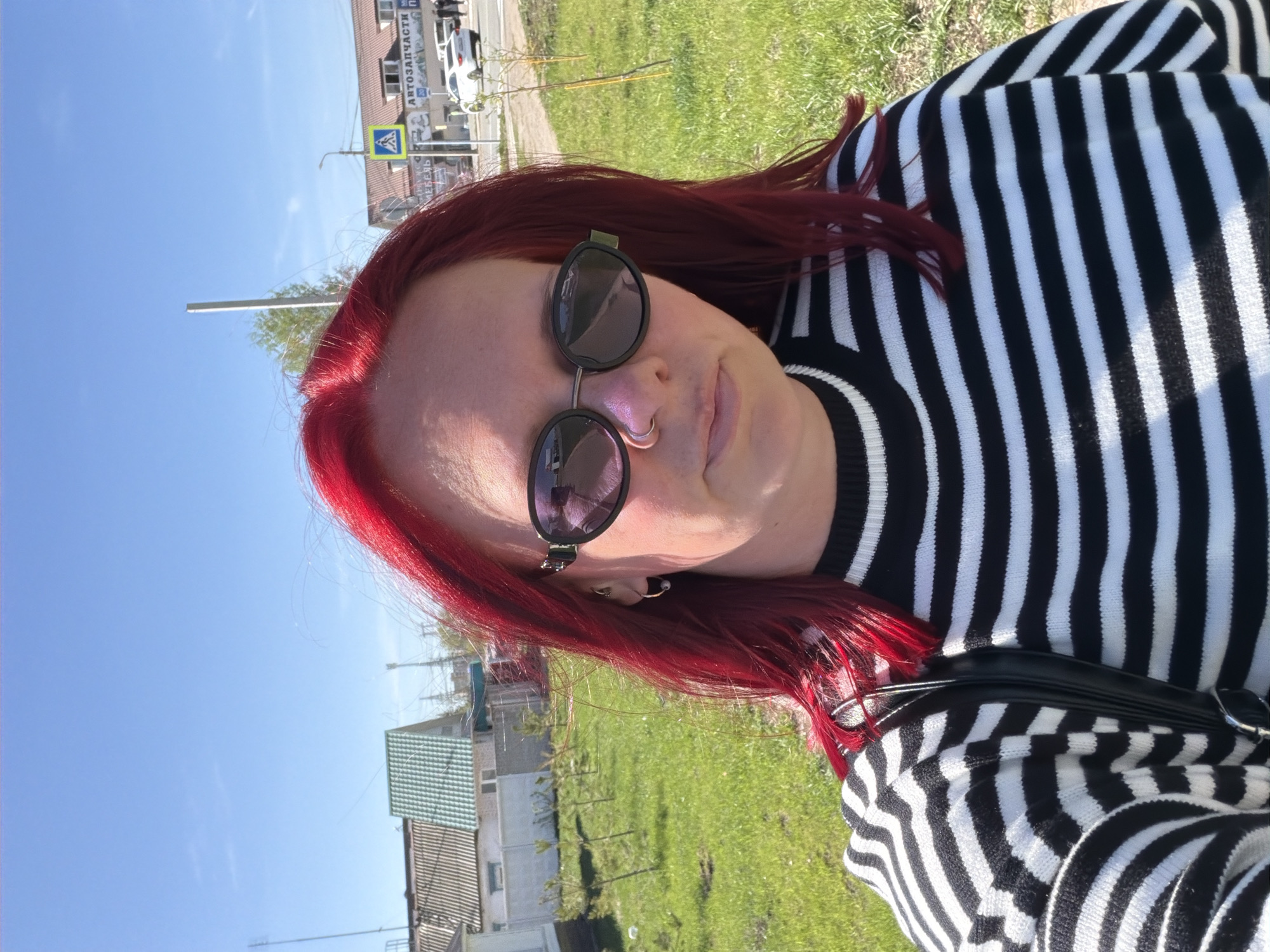 A person with red hair and round sunglasses is taking a selfie outdoors on a sunny day in what appears to be the city of Chaikovsky, Russia. They are wearing a black and white striped sweater and a nose ring. Buildings, grass, and a road with parked cars are visible in the background, along with a sign in Russian that reads "АВТОЗАПЧАСТИ" (Auto Parts) and a pedestrian crossing sign.