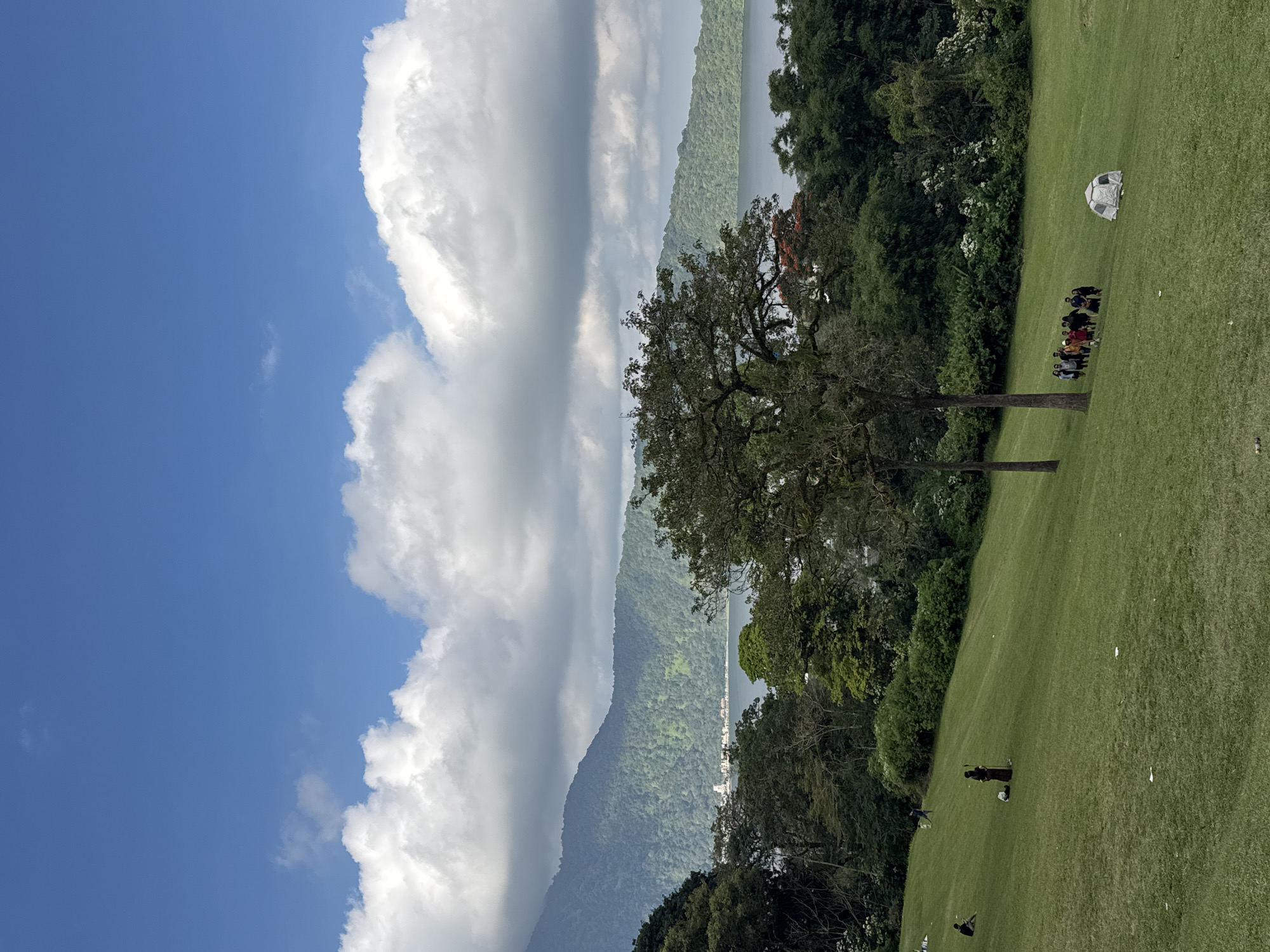The image captures a scenic view in Baturiti, Indonesia. The foreground is dominated by a lush green grassy field, which slopes gently downwards. Several people are scattered across the field. In the mid-ground, a group of about eight individuals is gathered, appearing to be engaged in a conversation or activity. A small, white, dome-shaped structure, possibly a tent or a shelter, is visible further up the slope, to the right of the group. A lone figure is seen walking on the field in the lower right portion of the image, and another person is visible further in the distance. Two tall trees with dark bark and full canopies stand prominently in the middle ground, framing the scene.
The background features a dense, verdant forest that climbs up the side of a large mountain. Beyond the mountain, a body of water, likely a lake, is visible, with hints of buildings or settlements along its shores. The sky is a vibrant blue, partially obscured by large, dramatic white clouds that add depth and texture to the scene. The lighting suggests it is daytime, likely late morning or early afternoon, with clear visibility. The overall impression is one of a peaceful, natural landscape.