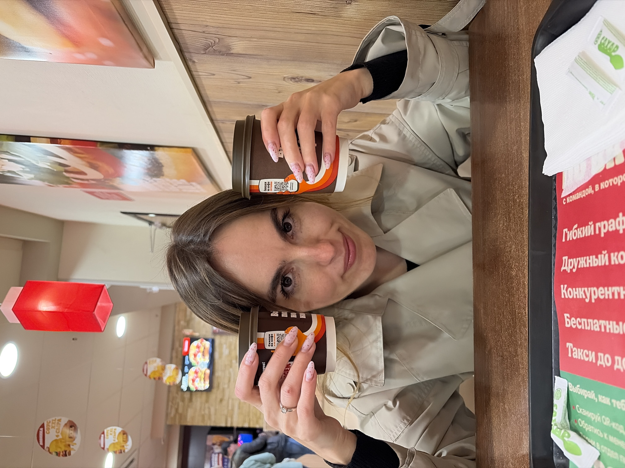 The image shows a woman indoors, holding two brown coffee cups. She is wearing a light beige trench coat over a black top. Her nails are decorated with a pink glitter design. She is smiling and looking at the camera.
Behind her, the interior of what appears to be a fast-food restaurant can be seen, including decorative elements, menu boards, and other customers. A red rectangular lighting fixture hangs from the ceiling. There is also a table to her right with sugar packets and text in Russian on it.
The location is likely a Burger King in Izhevsk, Russia, based on the interior design and promotional materials visible in the background.