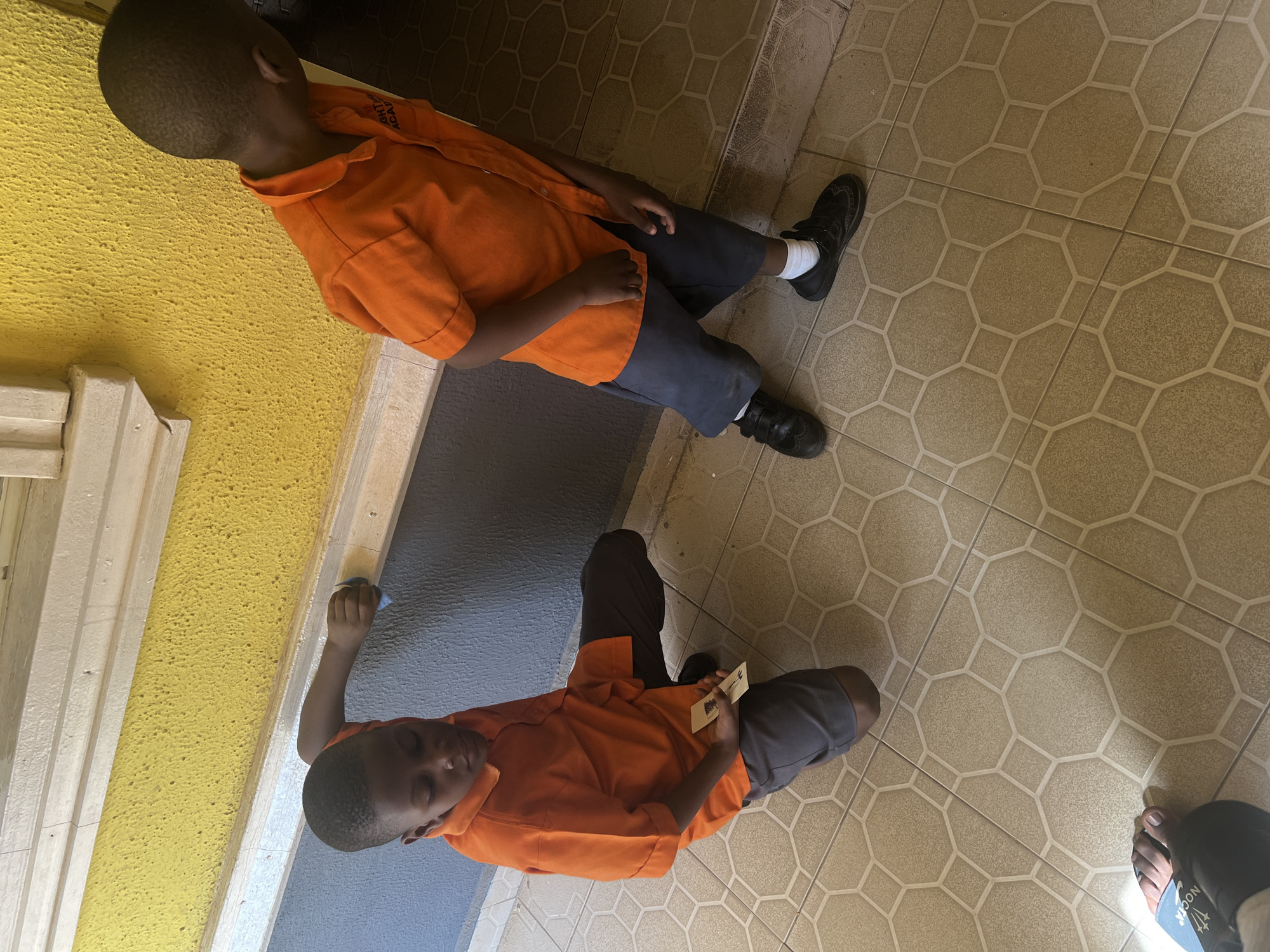 A young boy is sitting on a tiled floor in an unknown location in Markurdi, Nigeria. He is wearing an orange shirt and grey shorts, and black shoes with white socks. The wall in the background is painted yellow and blue. Another boy is sitting on the floor close to the first one. He is also wearing the same uniform, including orange shirt, grey shorts, and black shoes with white socks. The floor is tiled in a light brown geometric pattern.