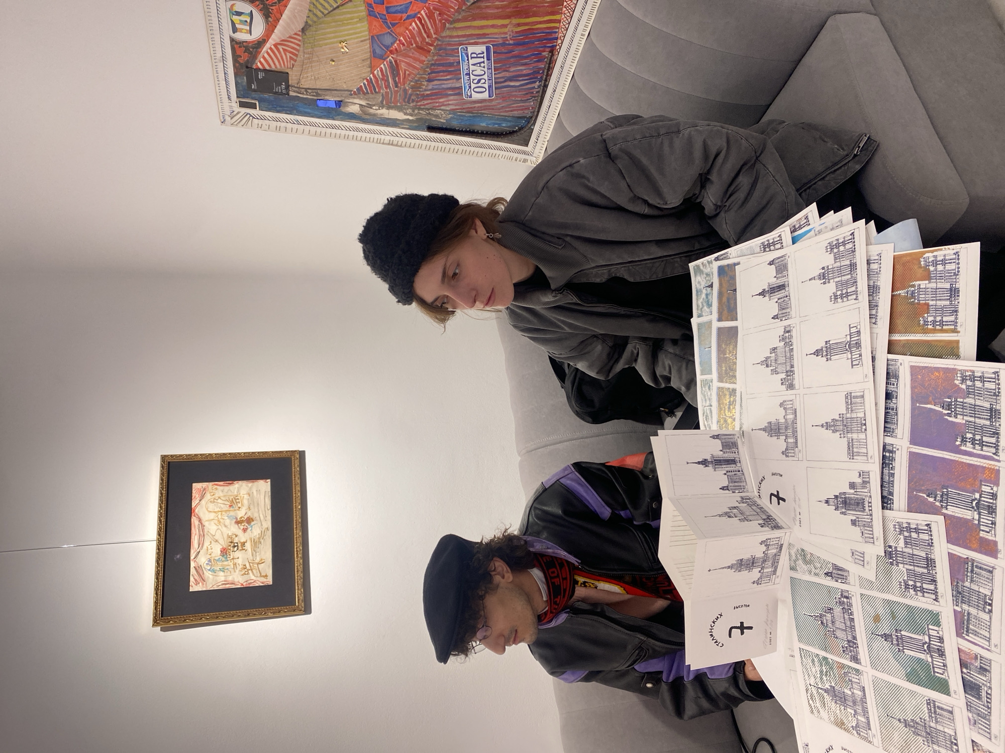 Two individuals are seated on a grey upholstered sofa in an indoor setting, likely an art gallery or studio. Both are intently focused on a collection of prints spread out in front of them, suggesting an art review or discussion.
The person on the right is a female wearing a dark, fuzzy beret and a dark grey padded jacket over a black top. She gazes downwards at the prints with a contemplative expression. Behind her, a large framed artwork is mounted on the wall, featuring an abstract or collage composition with geometric shapes and bold colors. Text visible on this artwork includes "OSCAR" and "NEW YORK" on what appears to be a license plate embedded within the piece.
The person on the left is a male, wearing a black flat cap, glasses, and a black leather jacket with purple shoulders, layered over a red and white patterned scarf. He is also looking down at the prints. To his left, on the white wall, a smaller, gold-framed artwork containing an abstract sketch is displayed.
The prints themselves are numerous and depict architectural sketches of large, ornate buildings. Many are monochromatic line drawings, while others incorporate various color washes or patterned backgrounds. Crucially, some prints clearly display the Cyrillic text "СТАЛИНСКИХ ВЫСОТОК" which translates to "STALINIST SKYSCRAPERS," indicating that the subjects are the iconic Stalinist-era high-rises found in Moscow. A stylized cross symbol resembling "†" is also visible on several sheets.
The scene conveys a focused, artistic atmosphere, likely a casual viewing or presentation of prints related to Moscow's architectural heritage. The artificial lighting evenly illuminates the subjects and the artwork.