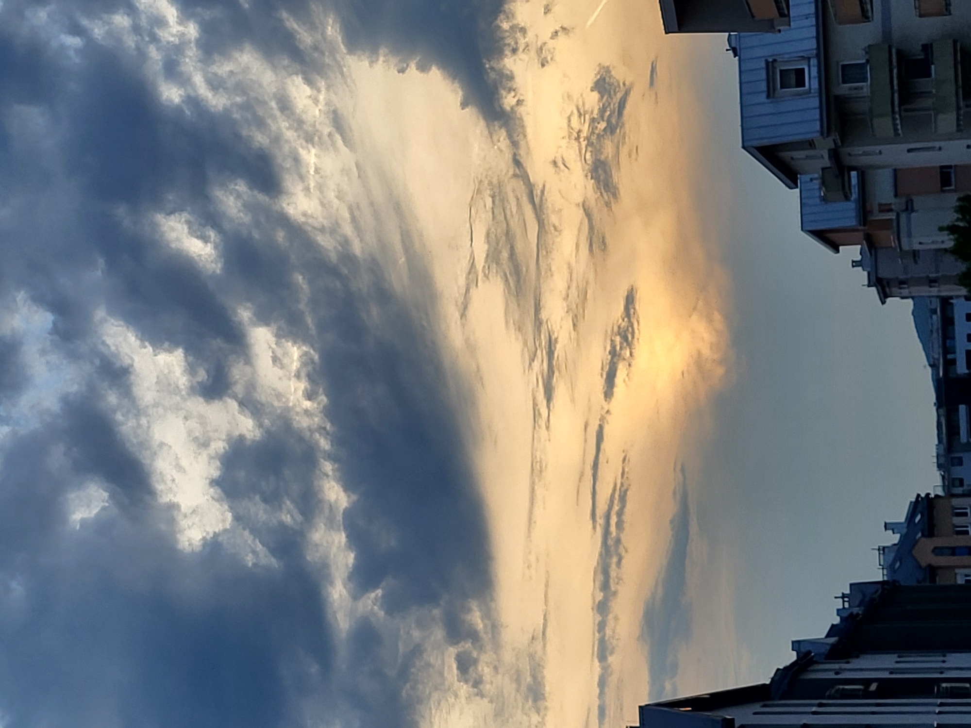 A vertical, low-angle, full shot captures the dramatic sky above buildings in an urban environment. The sky is filled with a mix of dark, heavy clouds and lighter, wispy formations, with a bright, glowing patch of sunlight near the center, suggesting either sunrise or sunset. The buildings in the foreground are silhouetted against the sky, with various architectural styles visible, including pitched roofs and balconies. The scene appears to be in a city in an unknown location.