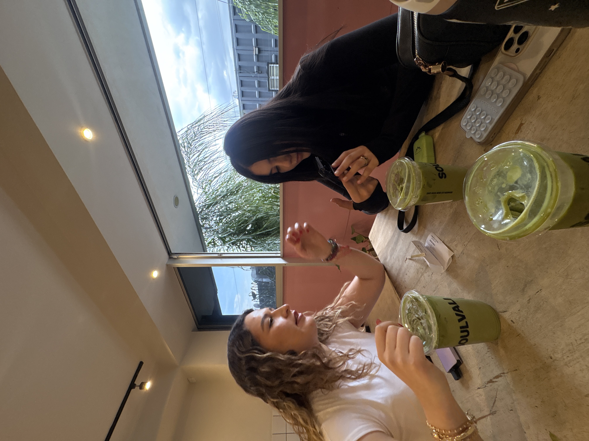 Two women are seated at a wooden table with two cups of green matcha drinks, likely in a cafe. One woman is wearing a black jacket and has long dark hair. She is holding a utensil and appears to be stirring or eating from one of the drinks. The other woman is wearing a white t-shirt and has curly blonde hair, gesticulating with her hand. The drinks appear to be from a brand called "Soulvalv." A phone case and a small black bag are also on the table. Outside the window, palm trees and urban buildings are visible. This location could possibly be in the city of Puebla, Mexico, given the architectural styles and vegetation visible.