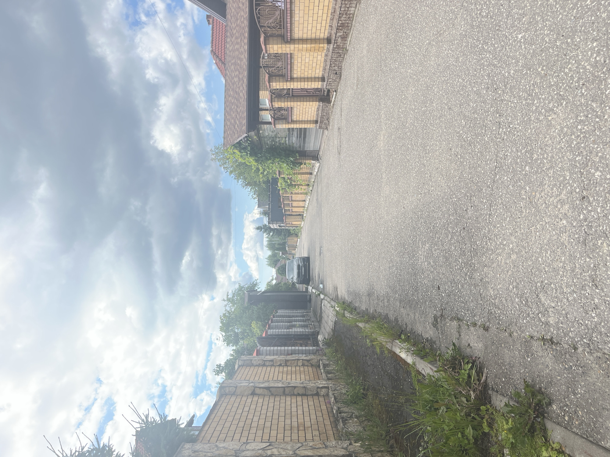 A street in Orekhovka, Russia, is shown from a street-level perspective. The asphalt road occupies the majority of the foreground and midground, with grassy areas and weeds growing along the sides. Brick fences with stone accents and decorative metalwork line both sides of the street, separating properties. Houses with tiled roofs are visible behind the fences. A dark-colored car is parked on the side of the road in the midground, and trees and foliage are present throughout the scene. The sky above is partly cloudy with a mix of gray clouds and patches of blue sky.