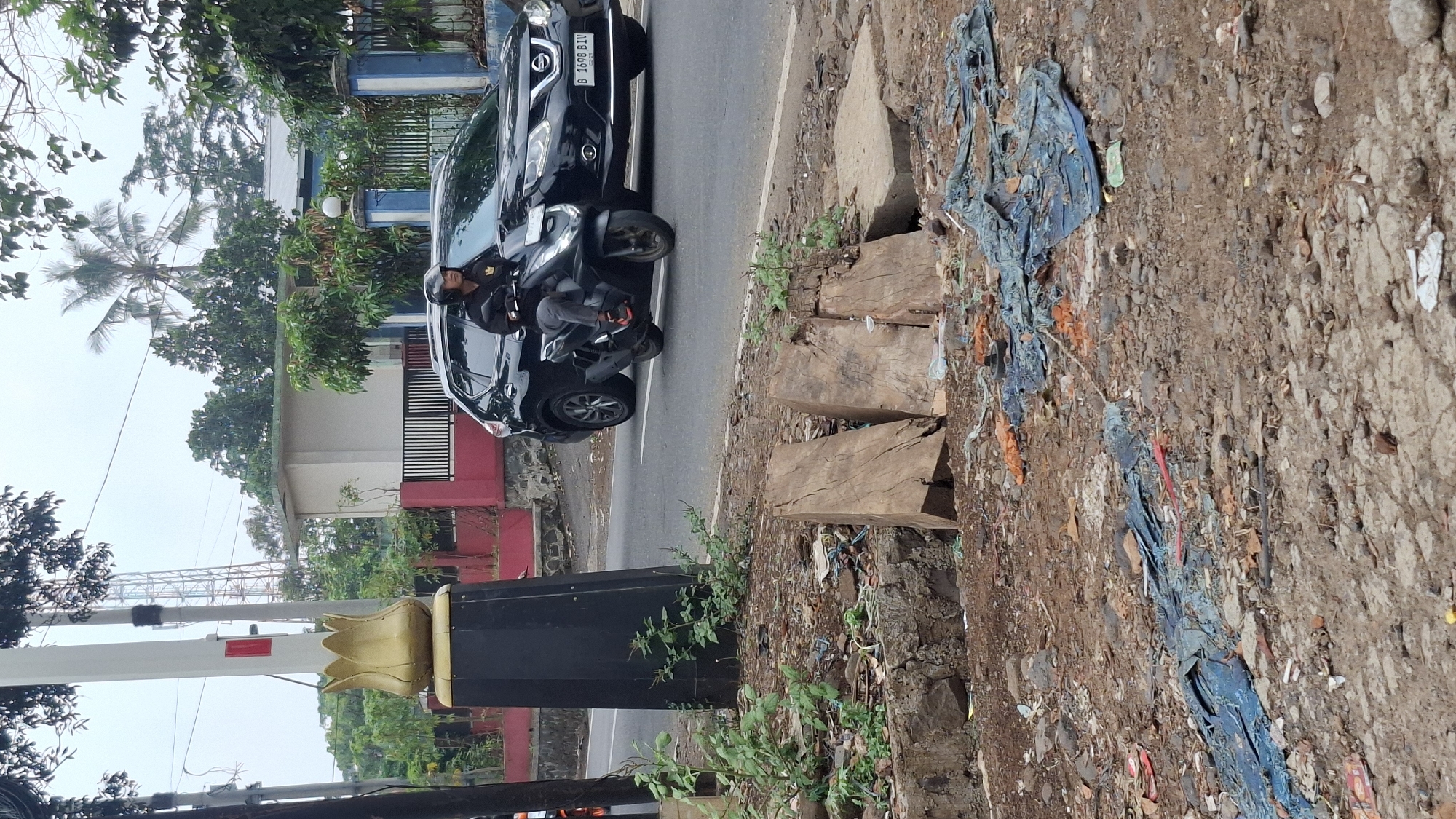 The image depicts a street scene in Cilaku, Indonesia, with a black SUV and a person riding a black scooter on the asphalt road. The scooter rider is wearing a helmet. In the foreground, there is a ditch filled with debris, including pieces of wood, plastic sheeting, and other trash. A black post with a gold tulip-shaped ornament stands to the left of the road. In the background, a building with a red trim and a fence can be seen, along with trees and a radio tower. The sky is overcast.