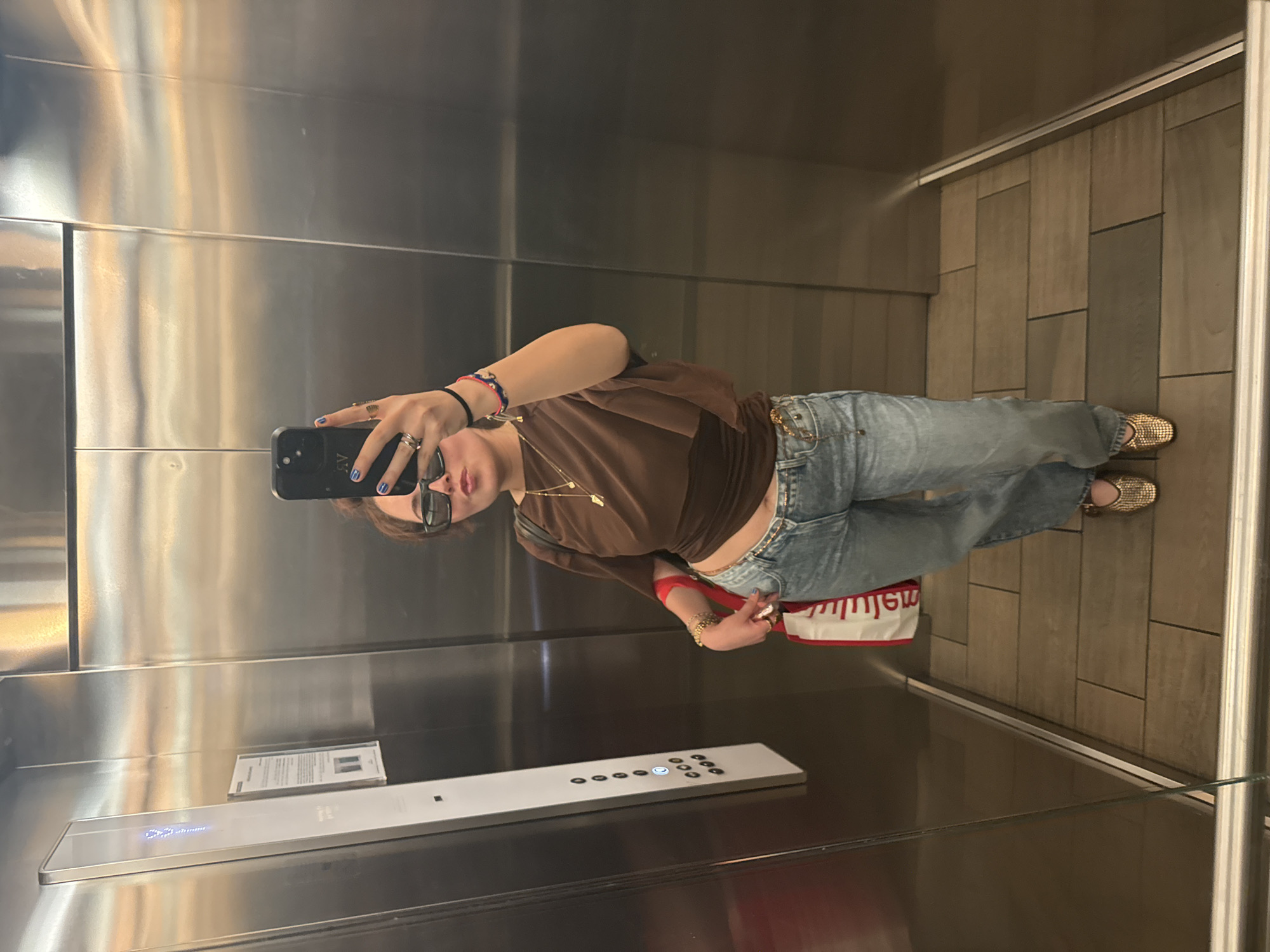 A young woman is taking a selfie in an elevator. She is wearing a brown shirt, blue jeans, and gold shoes. She has sunglasses on and is holding a black cell phone in her left hand. Her nails are painted blue. There is a red and white shopping bag near her right side. The elevator has a brushed metal interior and a panel of buttons. A paper on the elevator wall is visible. The location is likely in Mexico City, Mexico.