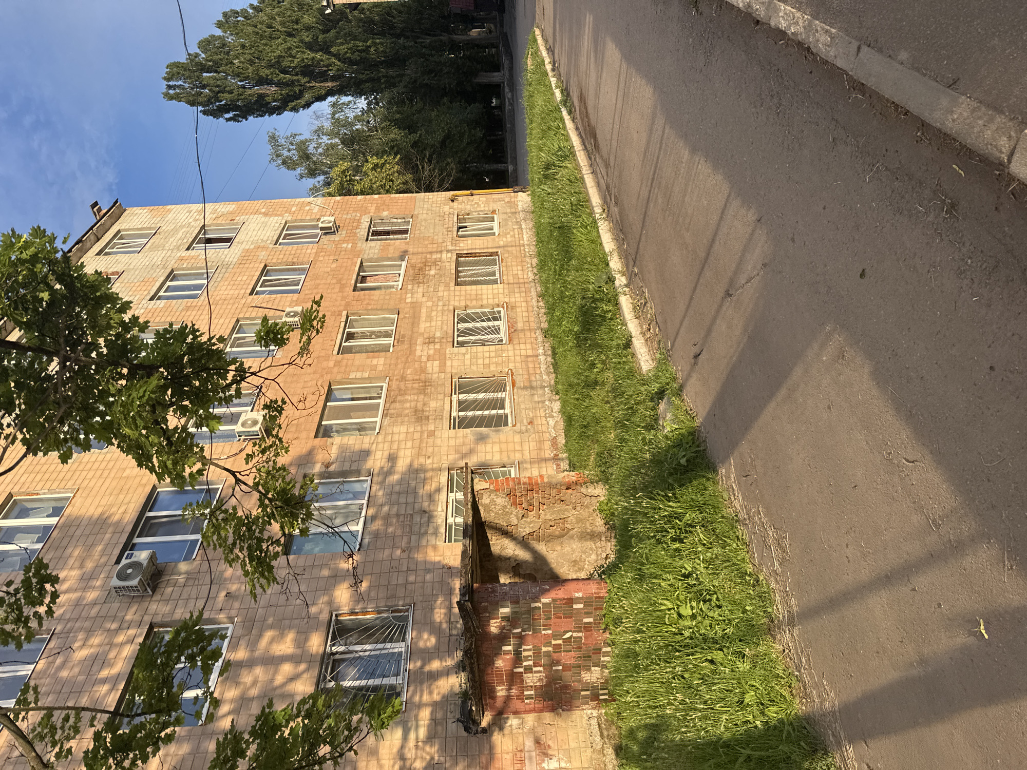 The image shows a segment of a multi-story brick apartment building with a tree in the foreground and a street in the foreground. The building has several windows, some with air conditioning units mounted outside. The brickwork is light brown and appears to be in good condition. There is a small, dilapidated structure with a red and white tiled facade attached to the side of the building at ground level, partially overgrown with grass. The street is paved and there is a strip of green grass between the street and the building. Tall, slender trees are visible in the background. The sky is blue with some clouds, and the sunlight casts long shadows across the scene, indicating it is likely late afternoon or early evening. No people are visible in the image.