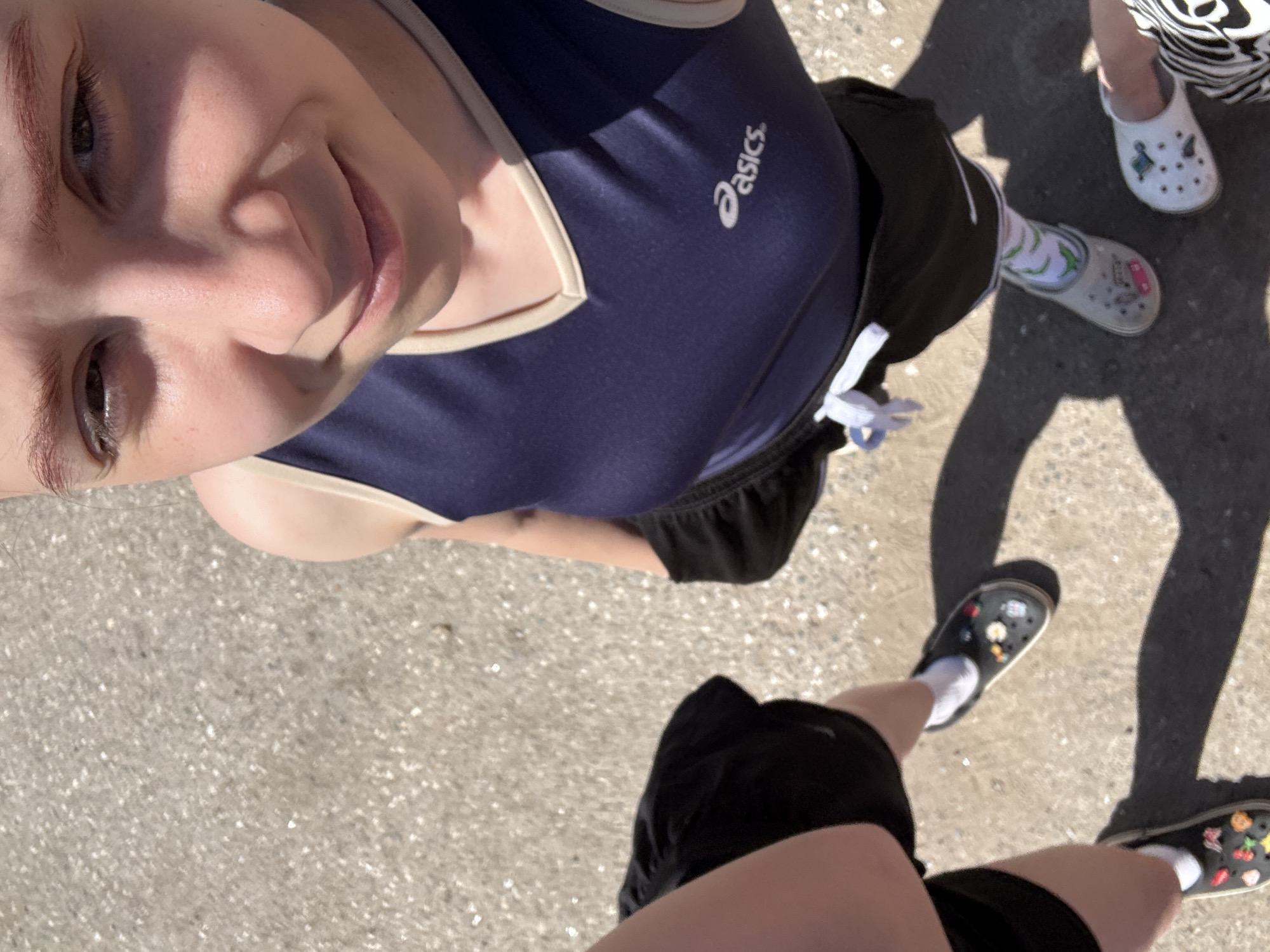 A high-angle, point-of-view photograph depicts a person wearing a navy blue athletic top with white trim and an "Asics" logo. The person is wearing black shorts and is standing on a concrete surface. In the lower right and upper right corners, are the feet of two other people wearing Crocs shoes. The background appears to be an outdoor setting, possibly a street or sidewalk. This image provides no location information.