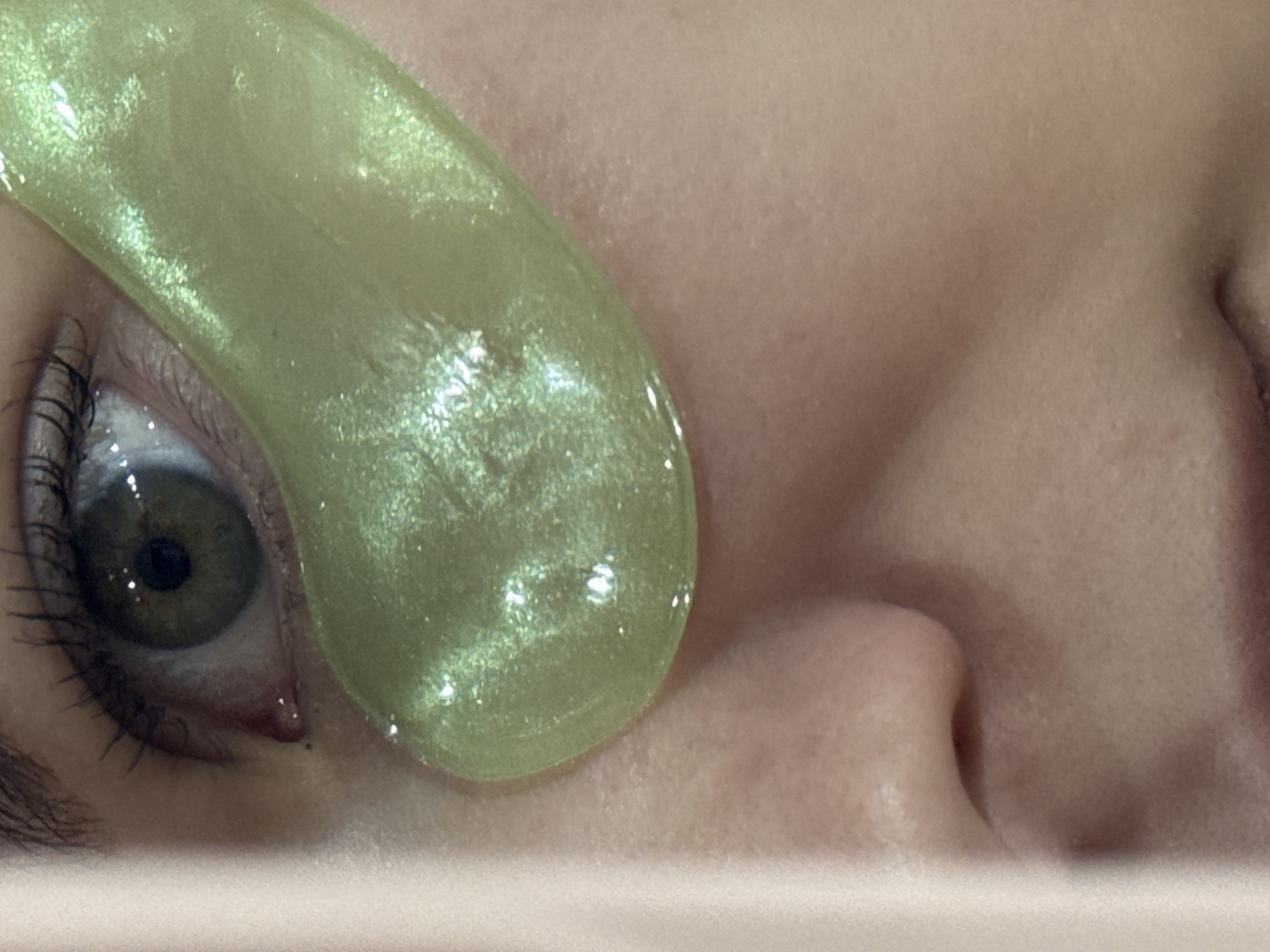 A close-up, indoor shot shows a person's eye with a green, shimmering under-eye mask applied. The person's eye is open, and the mask partially covers the lower portion of the eye and the skin beneath. The nose and part of the other eye, which is closed, are also visible. The person is lying on something light-colored, possibly a pillow or a surface. The location is not determinable from the visual information provided.