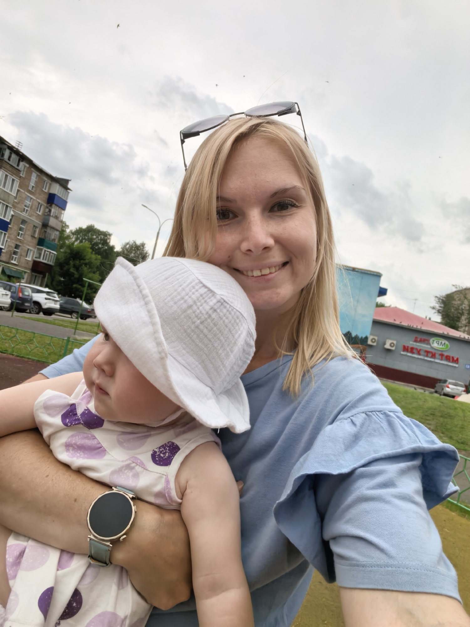 A selfie taken outdoors shows a blonde woman smiling, holding a baby wearing a white hat and a dress with purple polka dots. The woman has sunglasses perched on her head and is wearing a light blue ruffled shirt. A smartwatch with a grey band is visible on her wrist. In the background, there is a multi-story brick building, trees, a street with cars, a chain-link fence, and a building with a sign that reads "МАТРИ", which is consistent with the "Matri" supermarket chain found in Sterlitamak, Russia. The sky is overcast with clouds, and a few birds are visible flying. The setting appears to be a playground or park area.