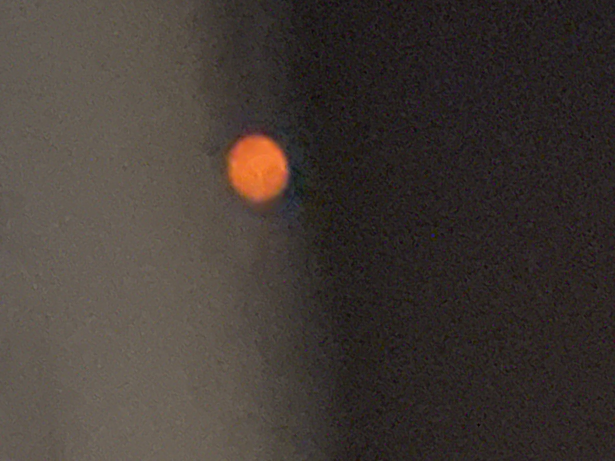 The image is a low-resolution, grainy photograph of an out-of-focus orange circle, possibly the sun or a light source, set against a dark, indistinct background. The background appears to be a gradient of muted grays and dark browns, suggesting a sky at dusk or dawn, or possibly a smoky or hazy atmosphere. There is no identifiable terrain, structures, or individuals in the image. The location is given as Stallings, United States of America, but the image itself provides no visual cues to confirm this.