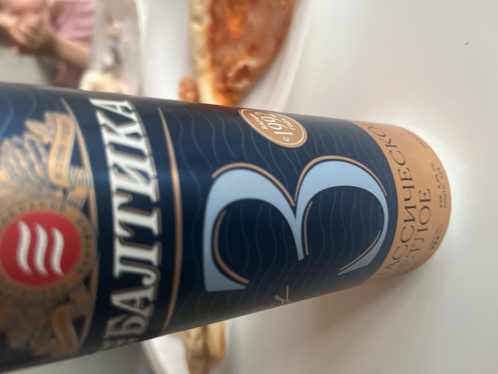 The image shows a close-up of a cup with the logo "Baltika" and the number "3" prominently displayed. The cup is dark blue with wavy patterns and gold accents. There is also a circular emblem with "1992" inside it. In the background, out of focus, appear to be plates of food, possibly pizza, and a person wearing a pink shirt. The lighting suggests an indoor setting, likely a restaurant or home. There is no identifiable information to place the image in Novocheboksarsk, Russia.