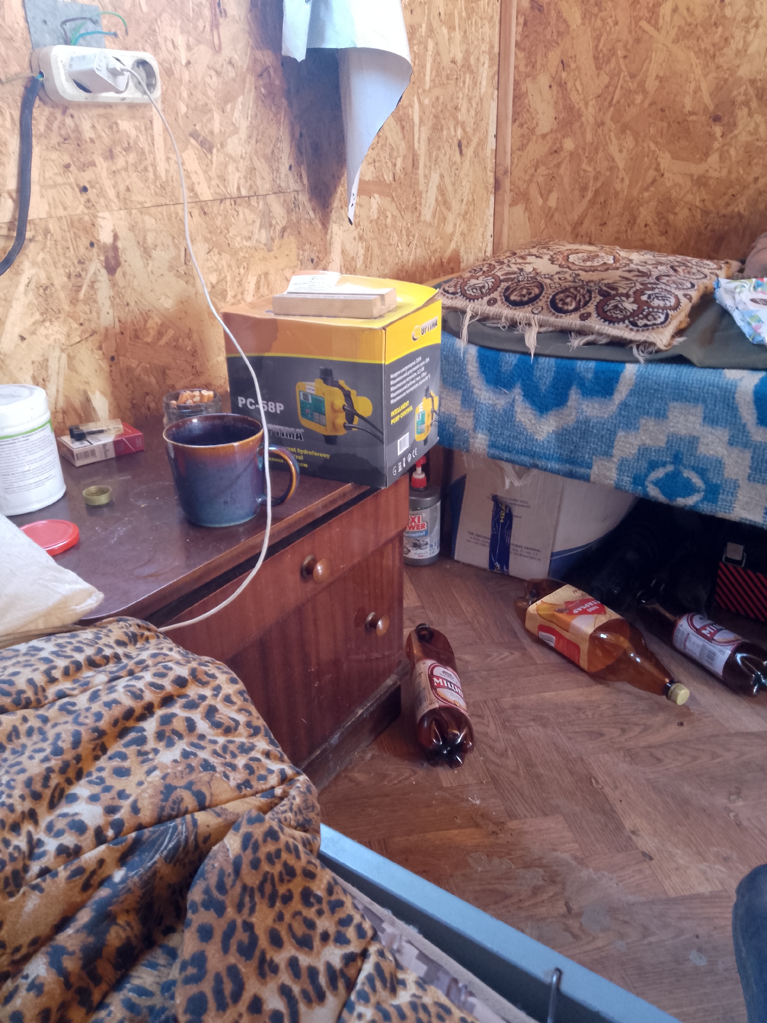 The image depicts a cramped, rustic room, possibly a temporary dwelling in Zalizne, Ukraine. The walls are constructed of particle board, and the floor is worn wooden planks.
In the foreground, a leopard-print blanket or mattress covers a surface, partially obscuring the floor. To the right, a blue metal frame is visible, suggesting a bed or bunk.
On a wooden dresser to the left, a white plastic container, a pack of cigarettes, a lighter, and a glass jar filled with cigarette butts are arranged. A dark blue and brown ceramic mug sits in the center of the dresser. A yellow and black box for a pump control system, labeled "PC-58P," is also present on the dresser, with its lid slightly ajar. A white cable runs from the wall socket to the box.
On the floor, several plastic and glass bottles are scattered. One is a large, brown plastic bottle with a label partially visible. Another is a glass bottle with a yellow cap. A smaller brown plastic bottle with a white label, and a partially visible glass bottle with a red label are also on the floor. A gray can with a red and white label, reading "XI power," is positioned near the dresser.
A blue and white patterned blanket is spread over a raised surface on the right side of the image, with a patterned pillow on top. The overall impression is one of a sparsely furnished, lived-in space.