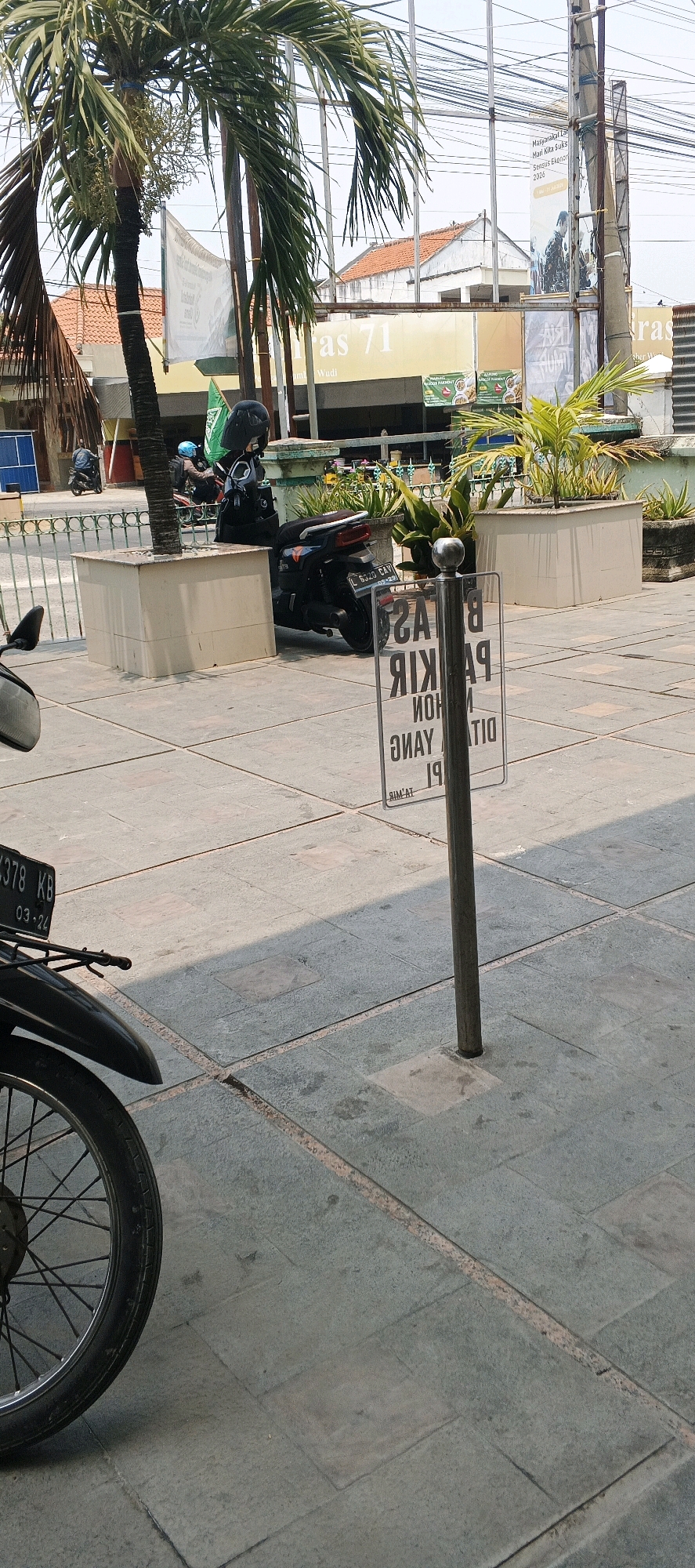 The image captures an outdoor scene in the city of Sumberwudi, Indonesia, during daylight. In the foreground, the front wheel and fender of a black motorcycle are visible on a tiled pavement. To the right, a metal pole supports a clear sign with text printed in reverse, which appears to be an inverted parking regulation sign. Behind the pole, a black scooter is parked, facing away from the viewer. Further in the background, a palm tree stands tall, its fronds casting shadows. Beyond the tree, a building with an orange tiled roof is partially visible, along with overhead electrical wires. A green flag is attached to a pole near the scooter. The environment suggests an urban setting with elements of nature and infrastructure.