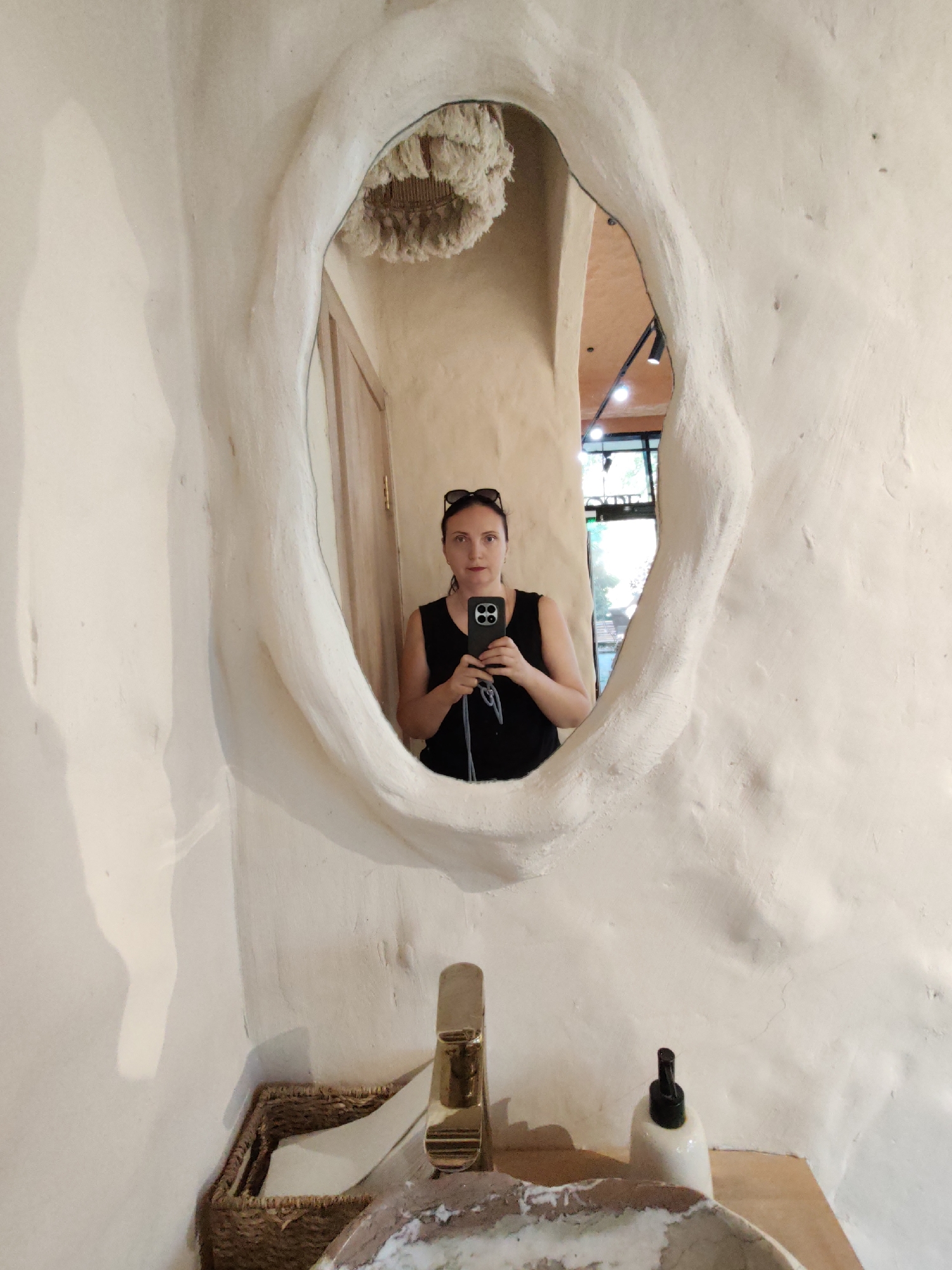 A woman takes a selfie in a mirror in what appears to be a bathroom. She is wearing a black sleeveless top and sunglasses pushed up on her head. She holds a black smartphone in her right hand, taking the picture. She is reflected in an irregularly shaped mirror with a textured, organic frame that is part of the wall. The mirror hangs above a sink with a gold faucet. To the left of the sink is a woven basket. To the right of the sink is a white soap dispenser with a black pump. The walls are rough and textured, painted a light off-white color. In the background, visible through the mirror, is a wooden door and a glimpse of a room with lighting fixtures. The style of the bathroom suggests a unique, possibly boutique, establishment, consistent with modern design trends found in tourist destinations. While the specific location within Sochi, Russia cannot be definitively determined from the image alone, the aesthetic is typical of contemporary cafes, hotels, or public restrooms designed with a focus on artistic and natural elements, which are common in popular resort cities like Sochi.