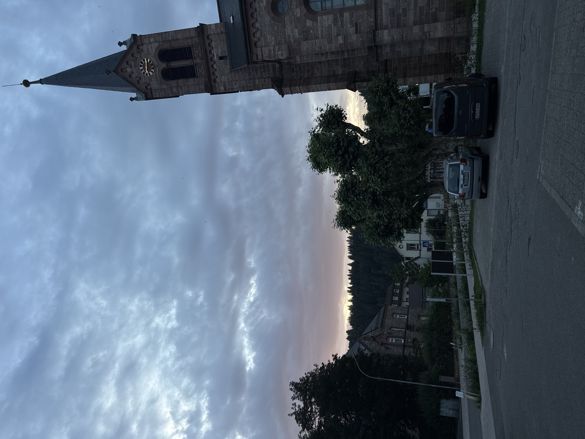 The image depicts a twilight scene in Schopfheim, Germany, with a prominent church steeple in the foreground. The sky is filled with dramatic, cloudy formations, transitioning from a pale blue to hints of pink near the horizon, suggesting the end of daylight. To the left of the church, a cluster of trees stands before a backdrop of a forested hill. Buildings, including what appears to be another older stone structure and modern white buildings, are visible nestled amongst the trees and along the road. A paved parking lot occupies the lower portion of the image, with two parked vehicles, a silver hatchback and a dark van. The overall atmosphere is serene and somewhat somber, indicative of the early evening.