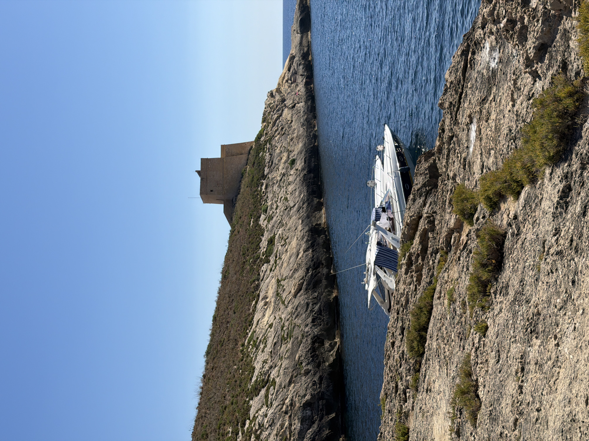 A sunny, clear day with a bright blue sky is depicted in Xlendi, Malta. The scene captures a coastal landscape featuring a rocky shoreline in the foreground, leading to the deep blue sea. On the shore, a white motorboat with blue and white striped coverings is moored. In the background, a rugged, vegetated hill rises, crowned by a historic stone tower, likely a watchtower, overlooking the water. The overall impression is one of a tranquil Mediterranean coastal view.