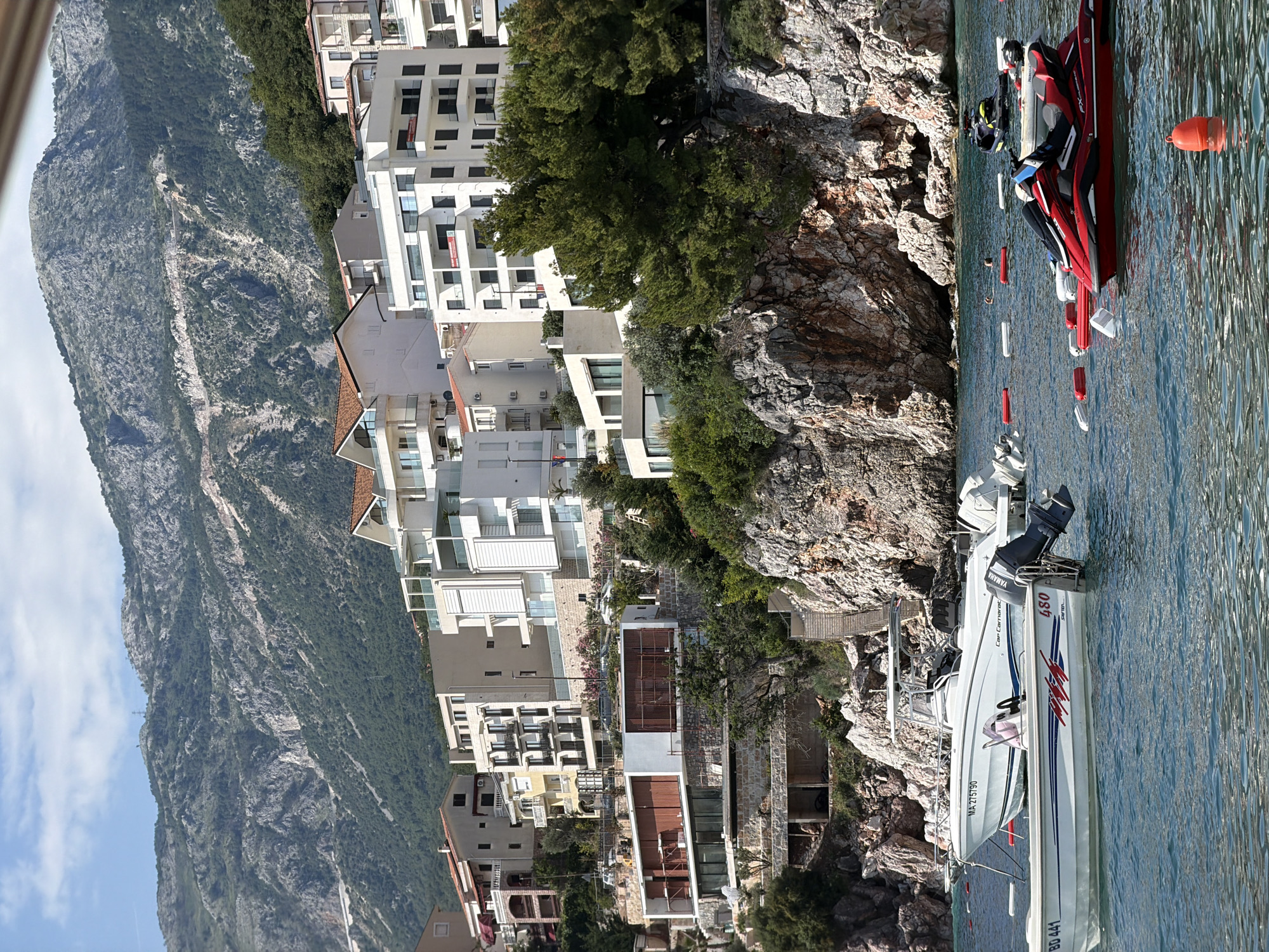 In Pržno, Montenegro, a coastal town nestled below a mountainous backdrop, the scene unfolds along a rocky shoreline dotted with modern white buildings. The azure water in the foreground is populated by various watercraft, including a white speedboat labeled "Car Camarat," and a smaller white boat with "BD 441" visible on its hull. A red jet ski, with a person seated on it, is positioned towards the right of the frame. Several buoys, both red and white, are scattered across the water's surface, marking designated areas. Behind the boats and the rocky cliffs, multi-story buildings with white facades and prominent balconies climb the hillside, interspersed with lush green trees. The imposing mountain range, characterized by its rugged, stony slopes and scattered vegetation, dominates the upper portion of the image, with a clear, partly cloudy sky above.