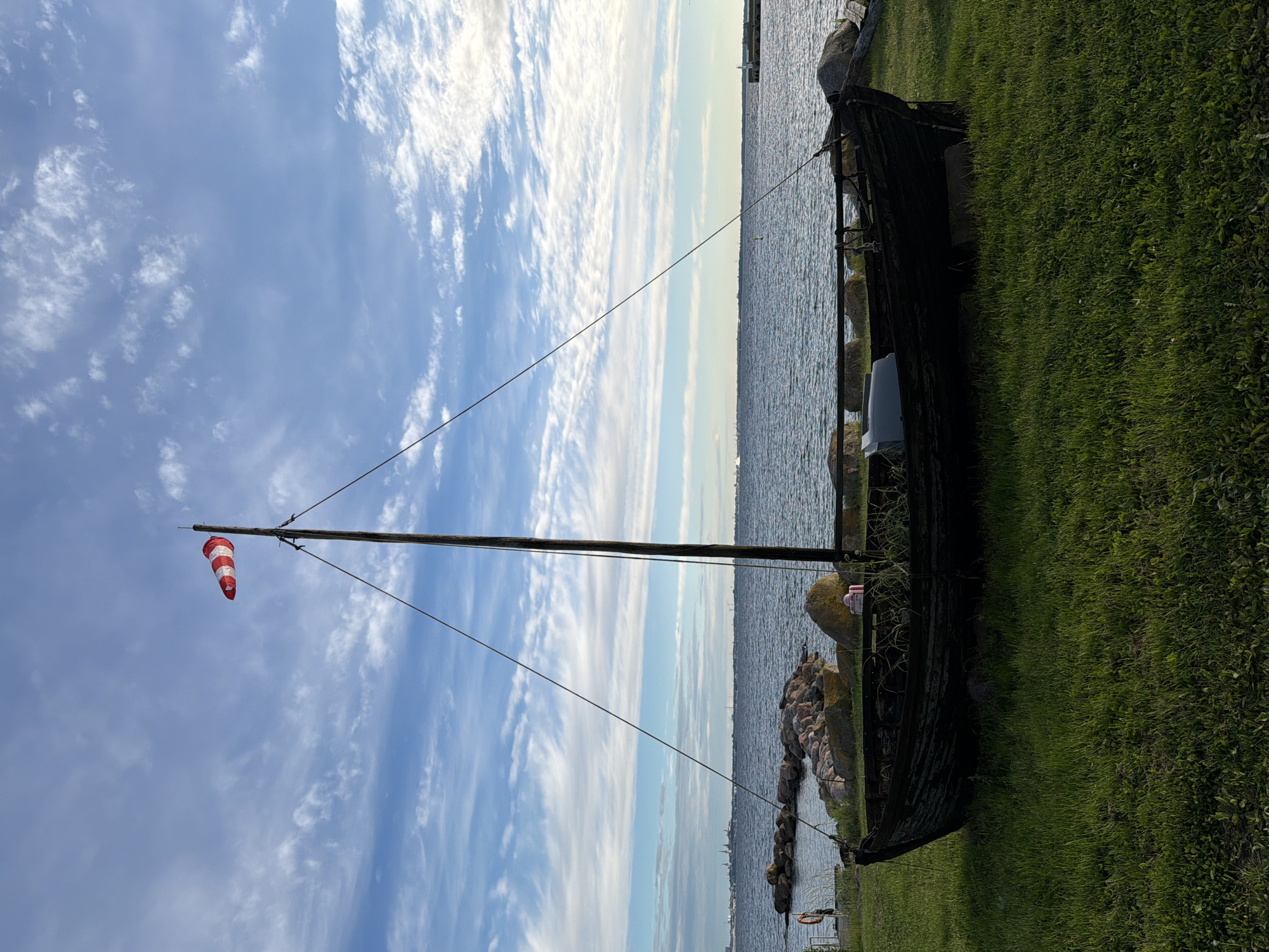 The image captures a scene in Viimsi, Estonia, featuring an old wooden boat resting on a grassy shore facing a body of water. A tall wooden mast, topped with a red and white striped windsock, stands erect from the boat. The boat is dark and weathered, with visible signs of age. Behind the boat, a rocky embankment leads down to the water, where the calm sea stretches towards a distant horizon. The sky above is a soft blue, adorned with wispy white clouds. In the distance, along the coastline, faint silhouettes of buildings and structures can be observed. The overall atmosphere of the image is serene and suggests a coastal setting.