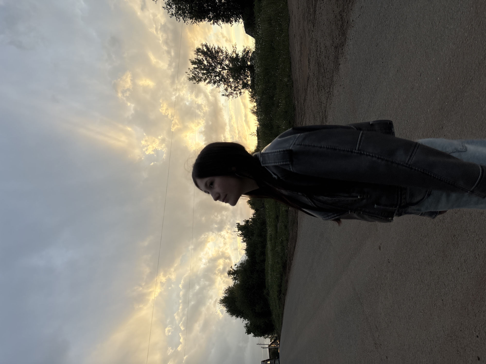 The image shows a young woman in Фоки, Russia, standing on a paved road. She is wearing a dark leather jacket and jeans. The sky is overcast with streaks of sunlight breaking through the clouds, and there are trees lining the side of the road.