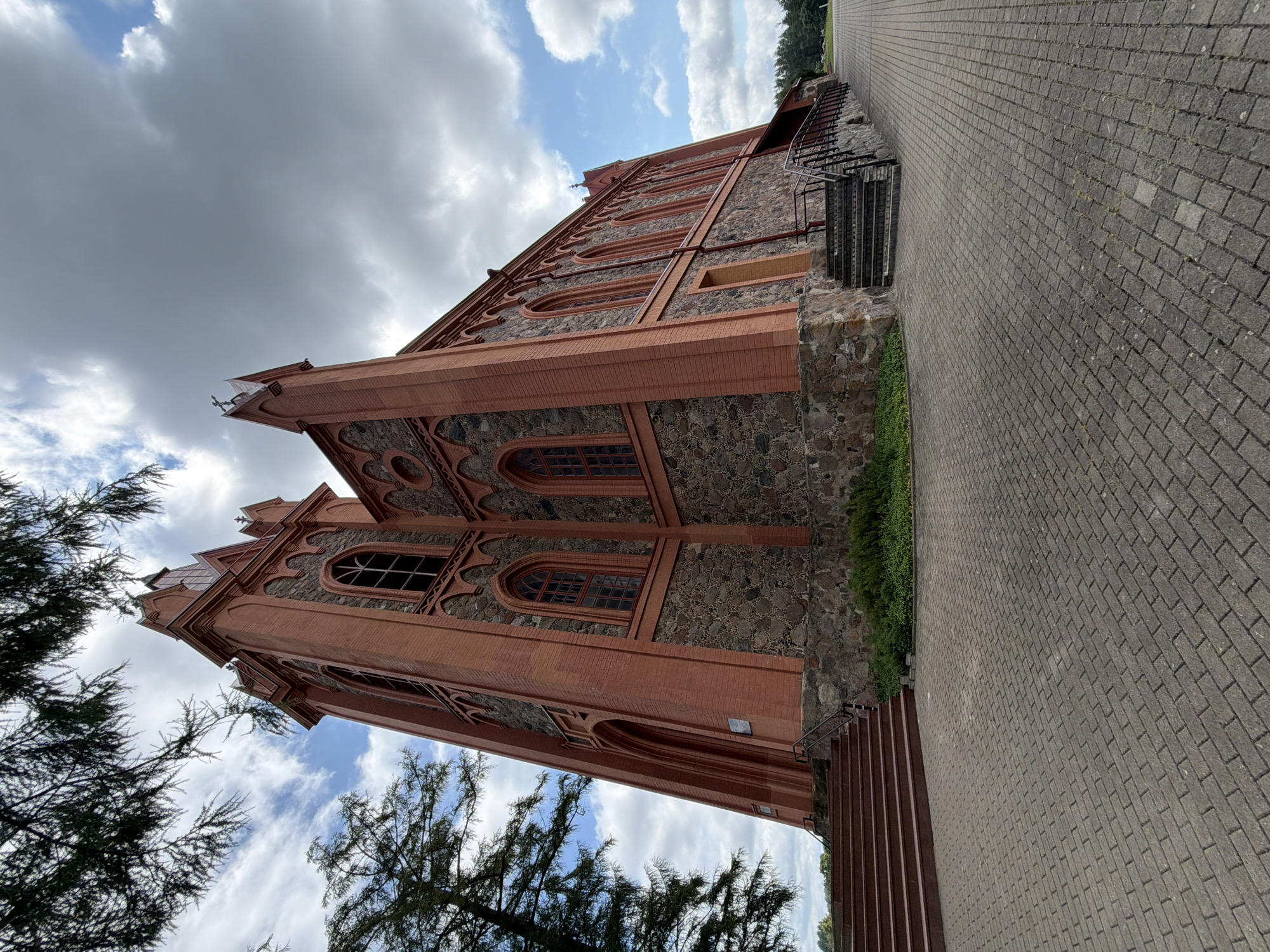 A low-angle, wide shot captures the exterior of a gothic-revival church in Dūkštos, Lithuania. The church is constructed with red brick and stone, featuring pointed arch windows and a tall, slender tower. The foreground is dominated by a paved walkway of interlocking bricks, which extends to the right and curves around the building. To the left, a set of wide, red-brown steps leads up to an arched entrance. Green foliage is visible at the base of the building and a dark green coniferous tree partially frames the left side of the image, with its branches extending into the upper left corner. The sky above is a mix of heavy gray clouds and patches of bright blue, with scattered white clouds. There are no people or animals visible in the scene.