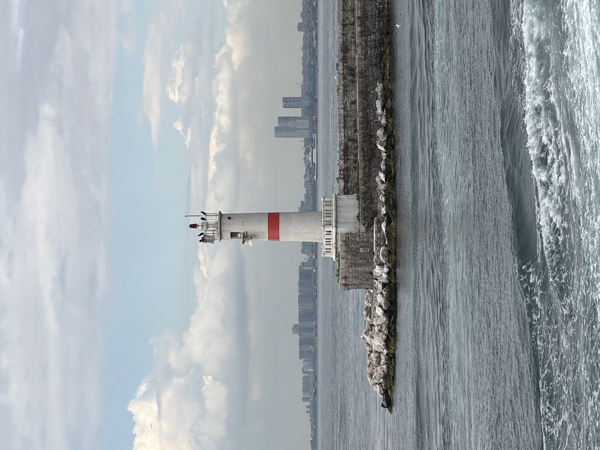 A lighthouse stands on a breakwater of stone and concrete in Kadıköy, Turkey. The lighthouse is white with a red band and has a red light at its top. Several people are visible near the top of the lighthouse. The sea is choppy, with waves breaking against the breakwater. In the background, the city skyline of Istanbul is visible across the water, with numerous buildings and skyscrapers. Clouds fill the sky, with an airplane visible in the upper left quadrant of the image.