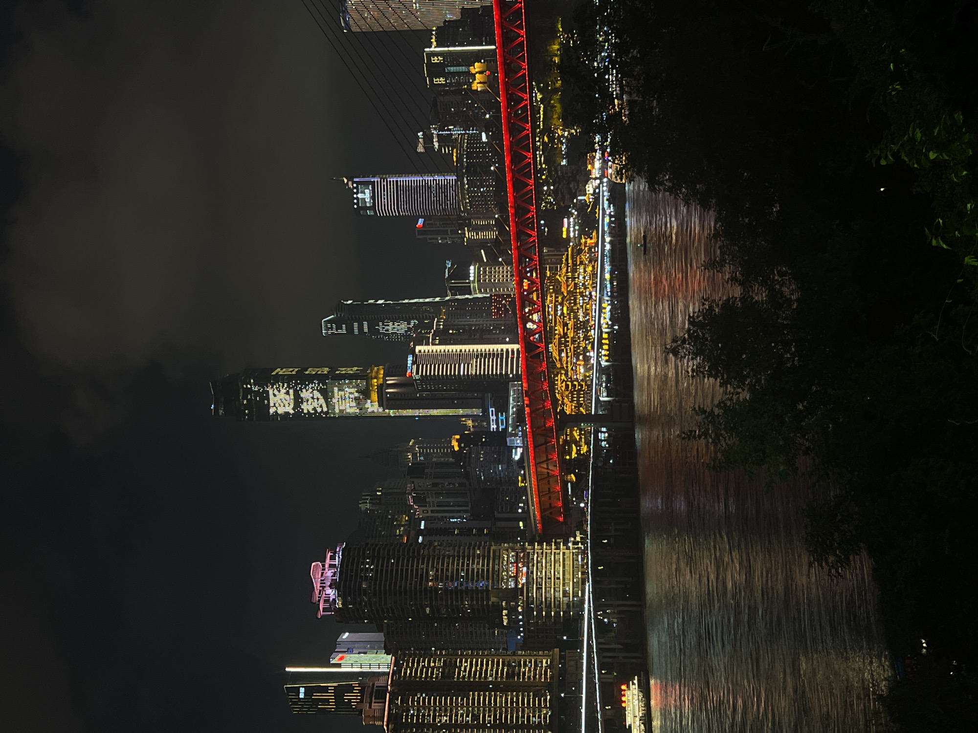 The image depicts a nighttime cityscape with a river in the foreground, reflecting the vibrant lights of a city skyline. A red illuminated bridge spans the river, connecting two parts of the city. Various skyscrapers of different heights and architectural styles dominate the background, many of them adorned with bright, colorful lights, indicating they are office buildings, hotels, or residential complexes. Several boats are visible on the river, some illuminated and moving, suggesting the river is a busy waterway. Lush dark green foliage in the foreground obscures the lower part of the view, creating a silhouette effect against the illuminated city. The sky is dark with some cloudy formations, typical of a nighttime scene. The overall impression is one of a modern, bustling metropolis at night, with a significant emphasis on illuminated architecture and infrastructure. The location appears to be 江北城街道, China, given the architectural style and the overall ambiance of a major Chinese city.