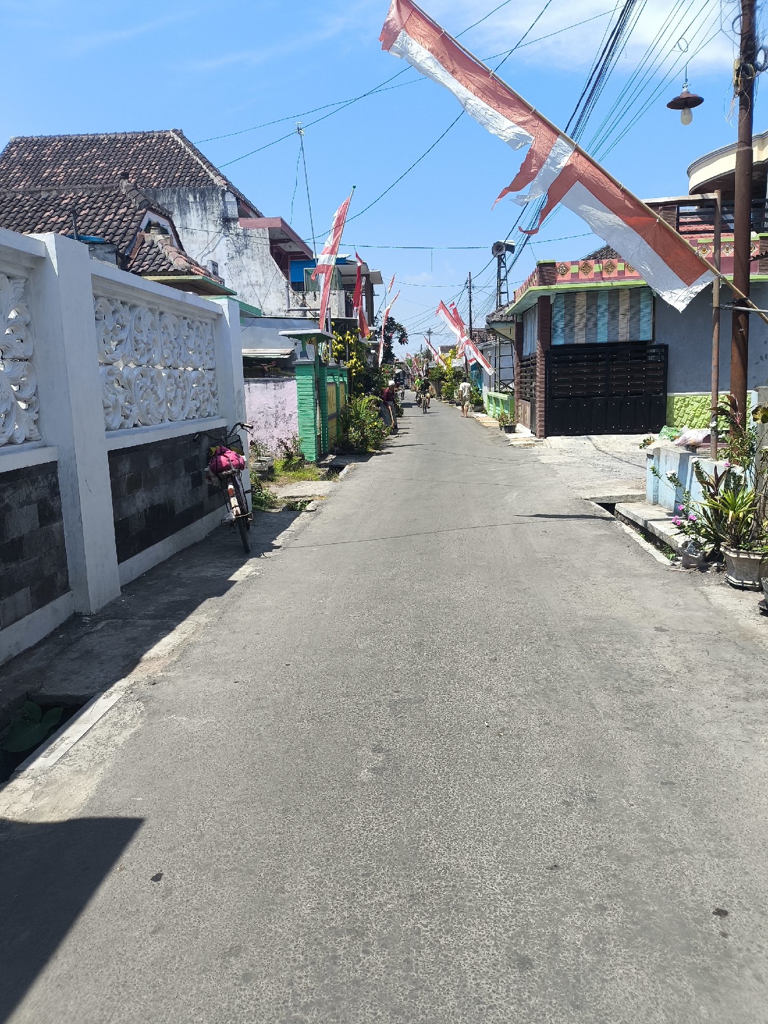 The image shows a street in Lumajang, Indonesia, decorated with Indonesian flags strung across the road. The street is paved and appears to be a residential area with houses on either side. A bicycle is parked on the left side of the street next to a white wall with decorative carvings. Several people are visible in the distance, some riding bicycles and others walking. The sky is clear and blue. The flags suggest a celebration or national holiday is taking place. The scene is characterized by the presence of Indonesian flags, typical residential architecture, and everyday street activity.