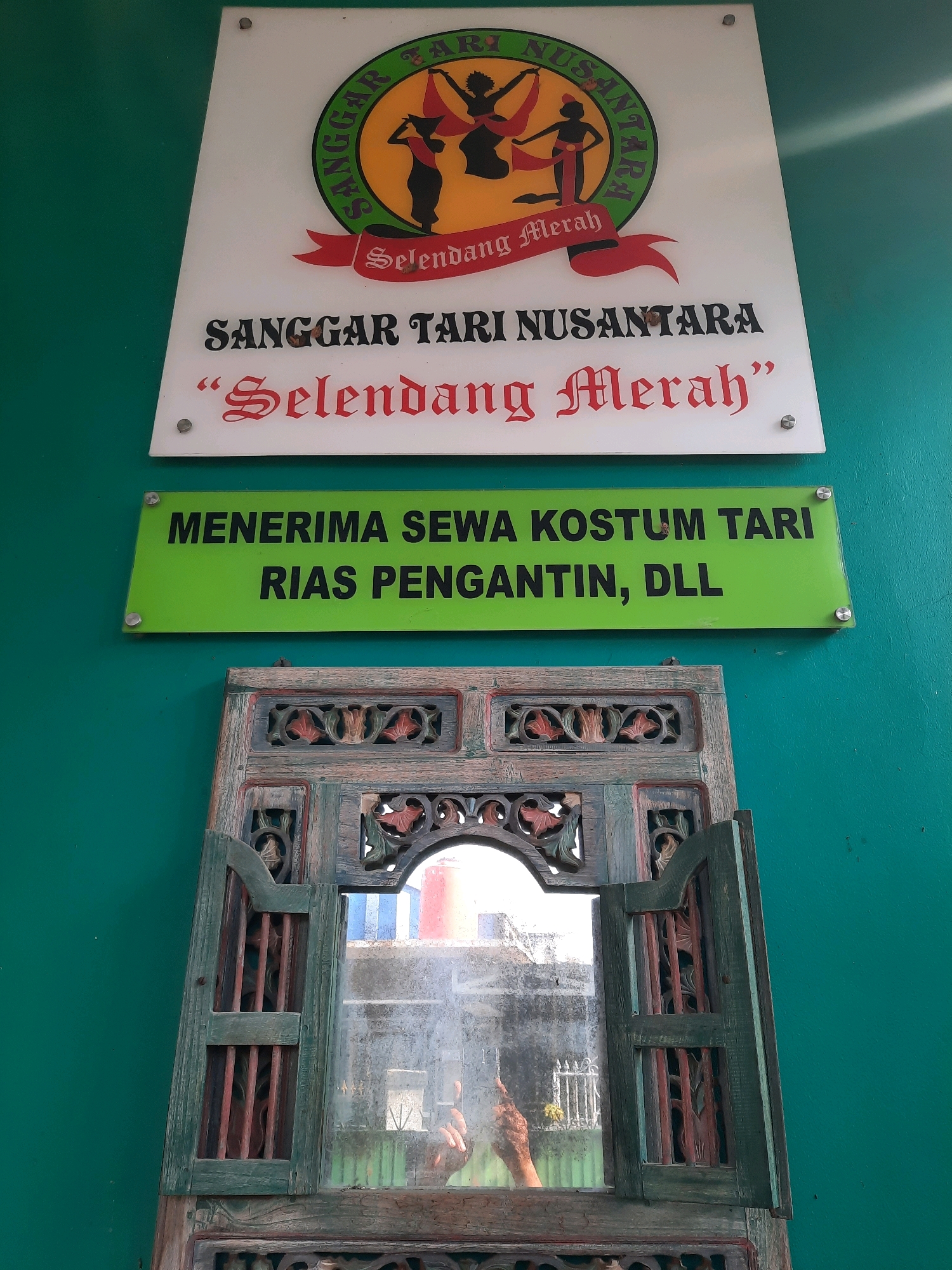 The image depicts a wall with signage and a decorative wooden mirror frame. The primary sign, at the top, is white with a circular logo that says "SANGGAR TARI NUSANTARA" and features silhouetted dancers. Below the logo, in a stylized script, it reads "Selendang Merah". Beneath this, in block letters, is "SANGGAR TARI NUSANTARA" followed by " 'Selendang Merah' " in the same script.
A second, rectangular green sign is below the first, displaying text in black. It reads "MENERIMA SEWA KOSTUM TARI RIAS PENGANTIN, DLL".
The bottom of the image features an ornate, antique-looking wooden mirror frame with intricate carvings and some distressed paint. The mirror itself is visible and reflects a somewhat hazy view of what appears to be a building, possibly with a water tank, and some fencing.
The background wall is painted a solid, vibrant teal green. There are small metal screws visible, securing the signs to the wall. The overall scene suggests an establishment related to dance and traditional attire, possibly a studio or rental shop. The location is identified as being in the city of East Jakarta, Indonesia.