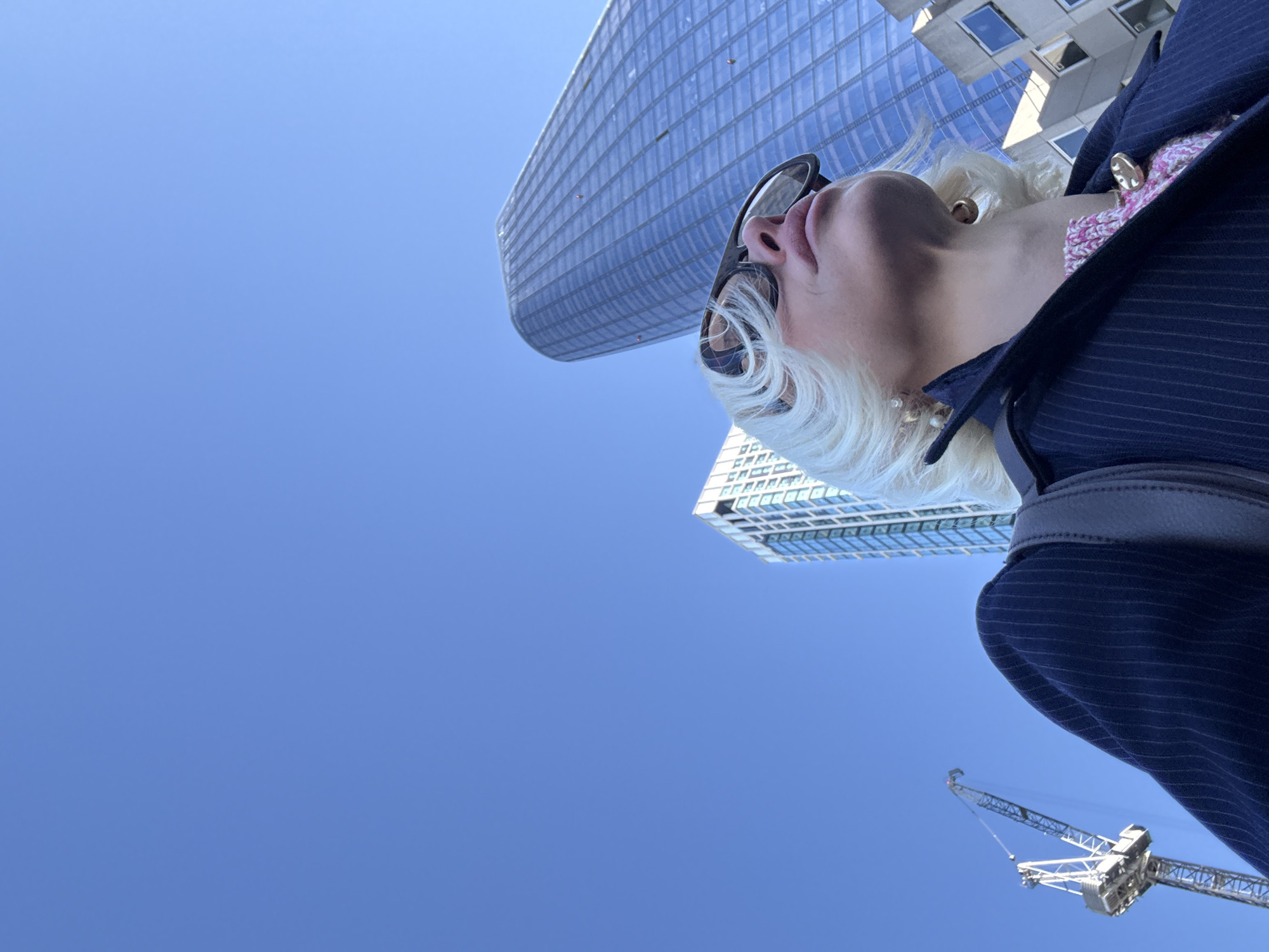 A low-angle outdoor image depicts an individual with blonde, wavy hair, wearing large, dark-framed sunglasses, a dark blue pinstriped jacket over a pink and white knitted top, and a gold circular pendant necklace. The person, positioned in the foreground, is looking upwards and to the right. Visible accessories include a gold hoop earring on the left ear, a pearl stud earring on the right, and a dark shoulder bag strap. In the background, the distinctive curved glass facade of 20 Fenchurch Street, known as The Walkie-Talkie, dominates the right side of the frame, reflecting a clear blue sky. To the left of this structure, a more rectangular glass skyscraper is visible, alongside a white lattice-boom construction crane. The scene is set against a clear blue sky in the City of London, United Kingdom.
