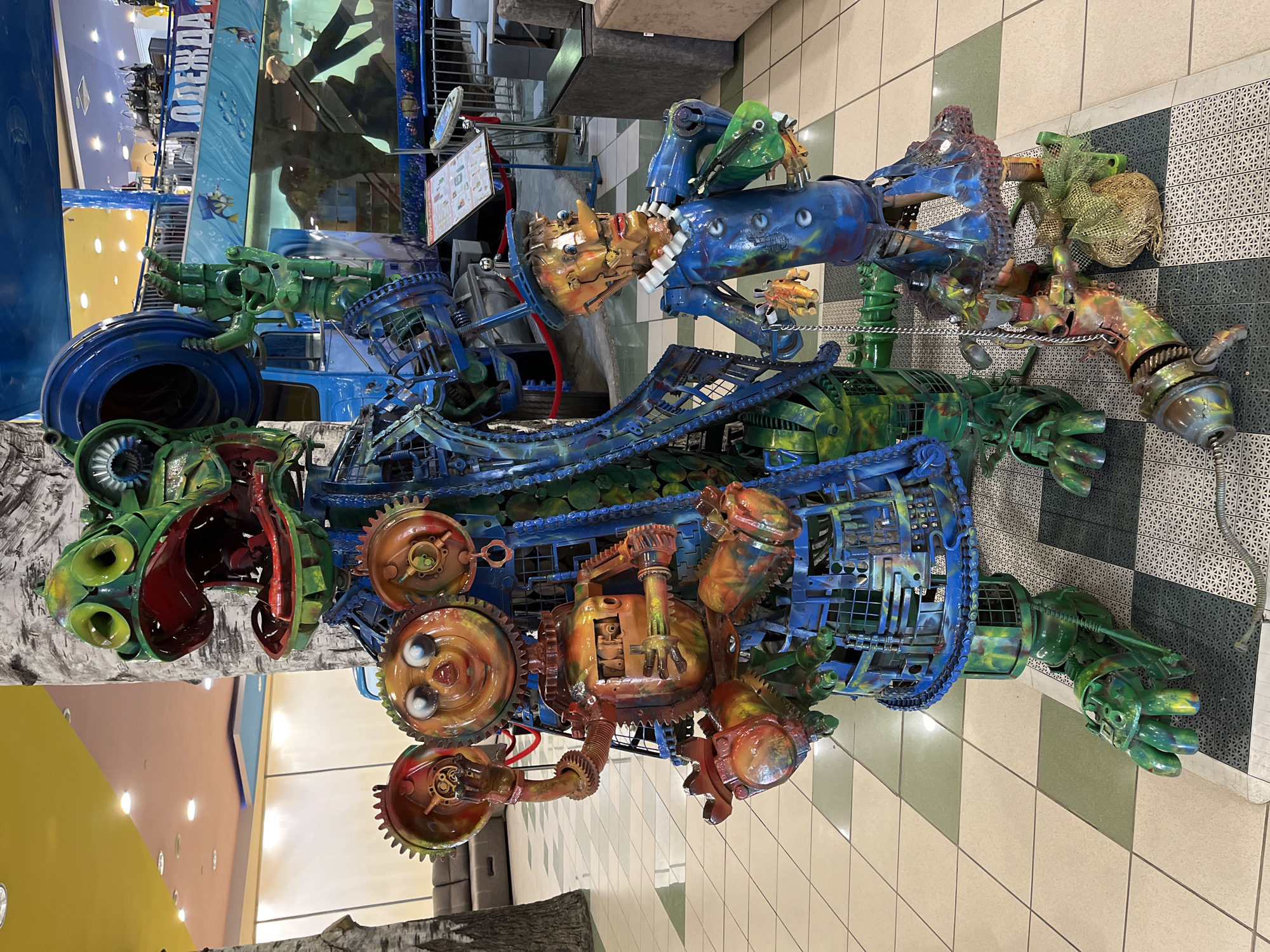 This media file features a large, colorful sculpture made of recycled materials, primarily metal. The sculpture is abstract and appears to be designed to resemble a fantastical creature or robot. It is positioned in what looks like an indoor public space, possibly a shopping mall or exhibition hall, given the tiled floor and the presence of a display with text and graphics in the background. The sculpture is predominantly blue, green, and orange, with various mechanical components such as gears, chains, and pipes incorporated into its design.
One section of the sculpture has a face-like structure with large, round eyes and a somewhat whimsical expression. Other parts resemble limbs with articulated fingers or claws. The overall impression is one of a whimsical, possibly steampunk-inspired creation. The lighting suggests it is daytime, but the indoor setting makes it difficult to determine the exact time.
In the background, there is signage, including a blue banner with white text that appears to be in Russian, reading "ОДЕЖДА" which translates to "CLOTHING". There is also a display with a chart or diagram and some other informational graphics. The setting suggests a commercial or recreational environment within Lyubertsy, Russia. The sculpture itself is the main subject, with no people actively interacting with it in the frame. The vibrant colors and intricate construction are notable details, suggesting it is a piece of public art or an exhibit.