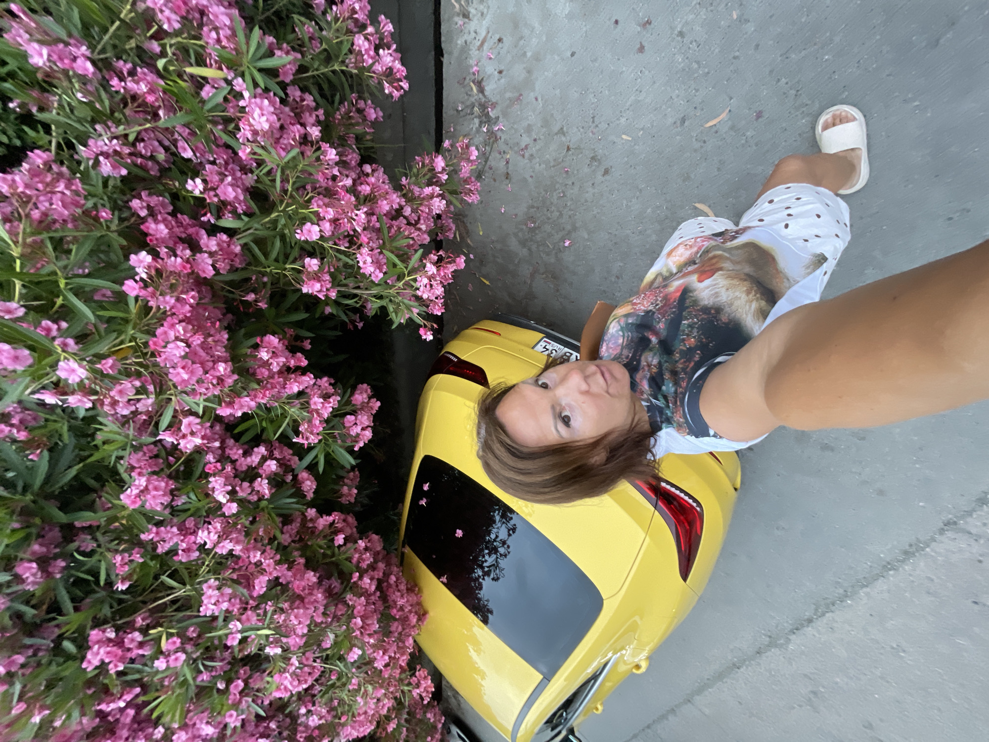 A woman with brown hair is taking a selfie outdoors in Sochi, Russia, with a yellow car and a bush of pink oleander flowers. The woman is wearing a t-shirt with a picture of an animal and a white skirt with cutouts. She is wearing white slides. The car is parked next to the bush of flowers and appears to be a modern sedan. The ground is paved and appears to be wet, suggesting recent rain. The lighting suggests it is daytime.