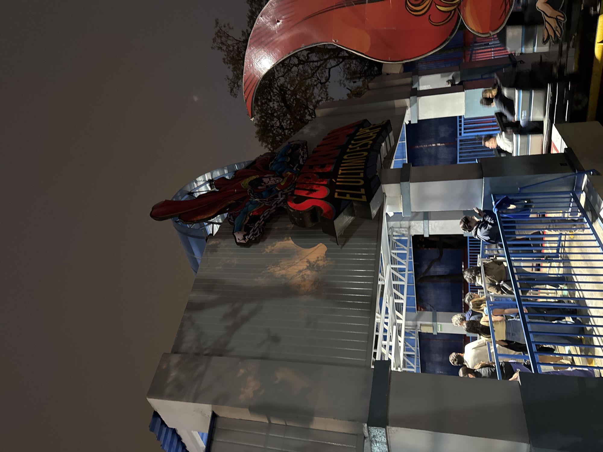 The image depicts the entrance to the Superman El Ultimo Escape roller coaster at Six Flags Mexico in Mexico City, Mexico. The entrance features a large Superman logo and sign. A queue of people waits on a staircase leading to the ride. Some people are seated on the roller coaster, ready to depart. The sky is overcast, and it appears to be dusk or night.