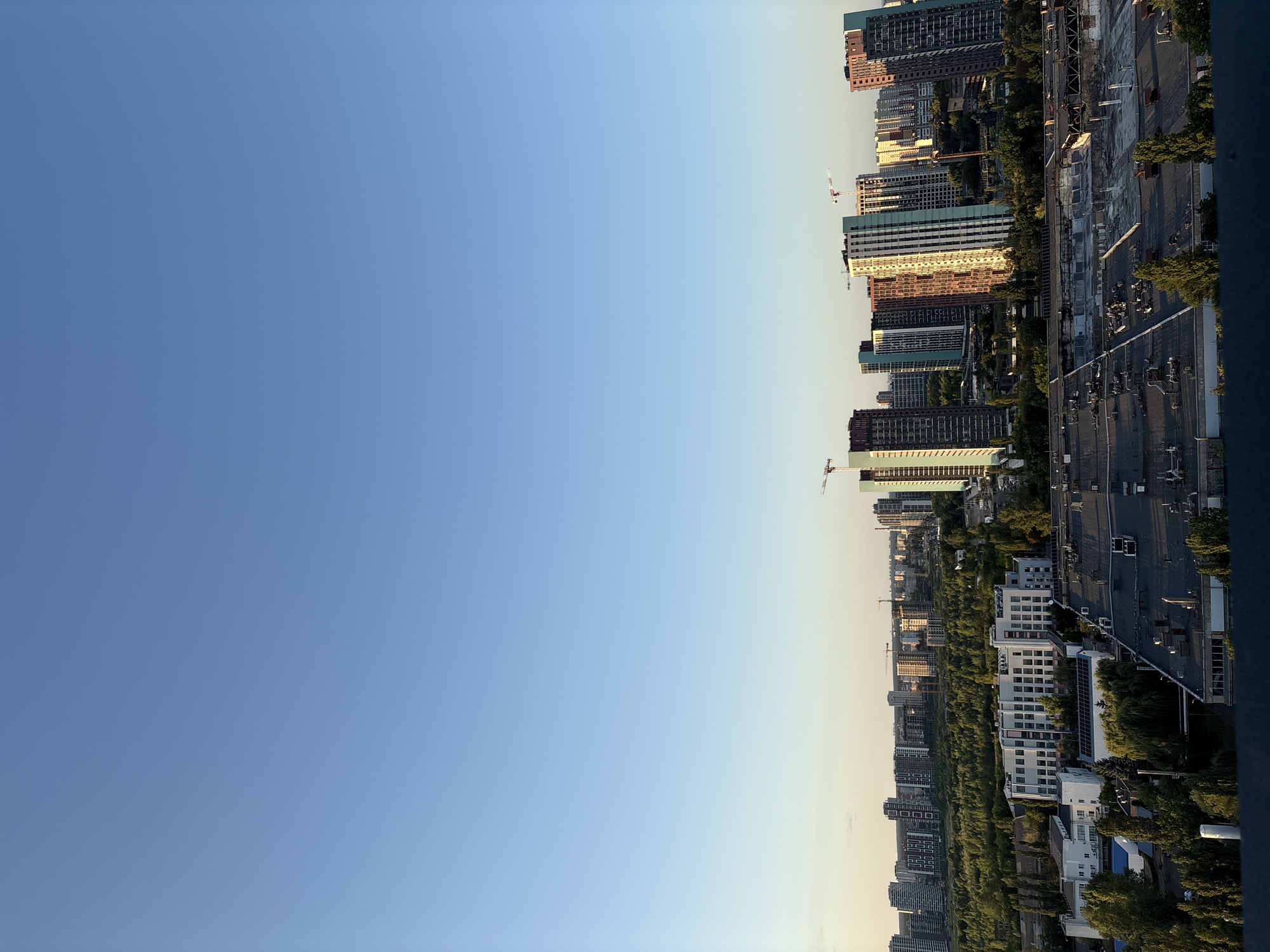 The image depicts an aerial view of a cityscape in Kyiv, Ukraine, under a clear blue sky. The foreground is dominated by the flat, dark rooftops of industrial or commercial buildings, some with ventilation units and skylights visible. Beyond these structures lies a dense green forest, separating the industrial area from a cluster of high-rise residential buildings. These apartment buildings vary in height and architectural style, with some featuring colorful facades, suggesting a relatively modern development. Several construction cranes are visible among the buildings, indicating ongoing development in the area. The overall scene captures a blend of industrial infrastructure, natural greenery, and modern urban housing, typical of a growing metropolis. The lighting suggests it is late afternoon or early evening, with the sun casting long shadows and illuminating the facades of some of the taller buildings.