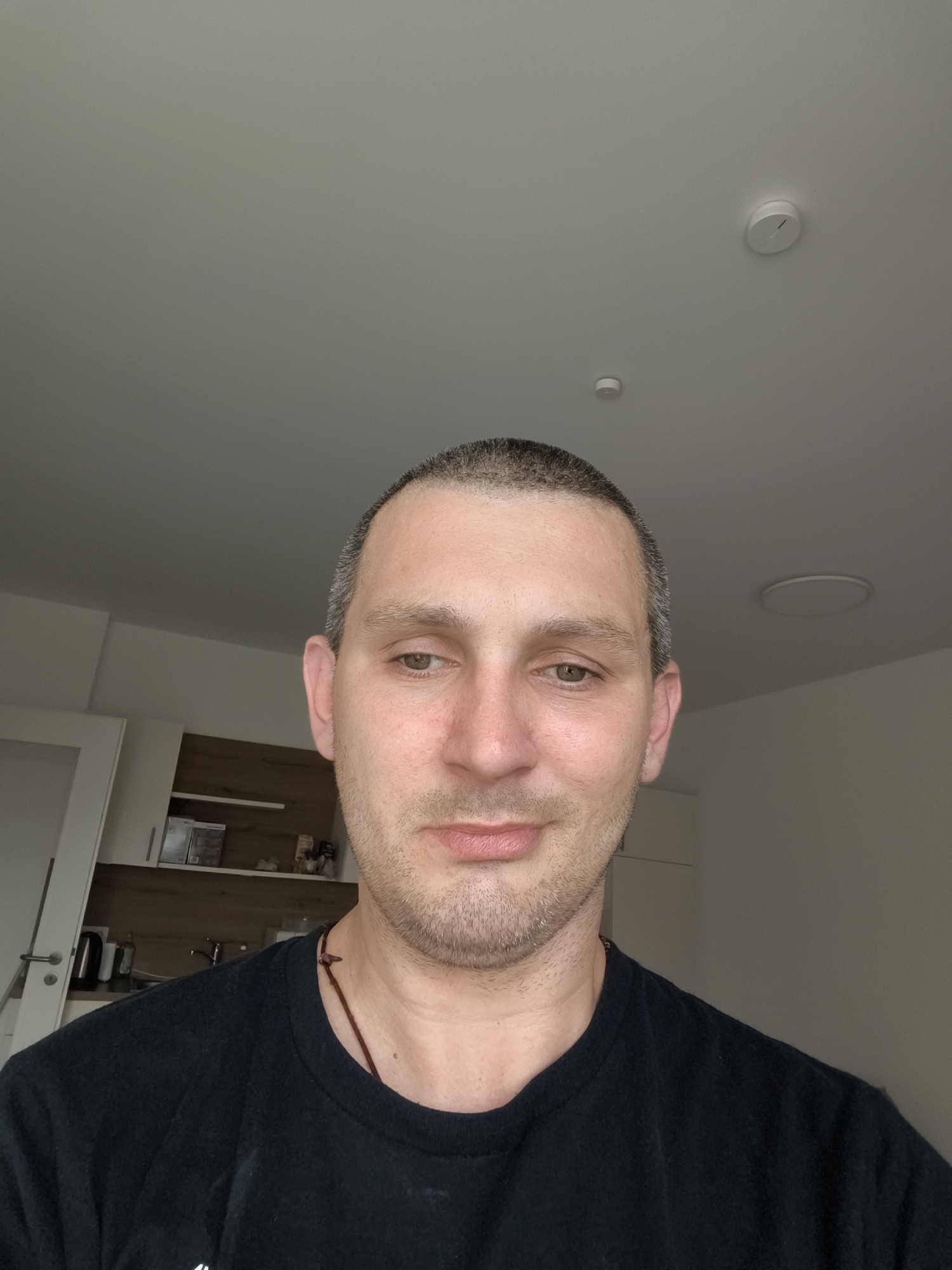 The image shows a medium shot of a Caucasian male wearing a black t-shirt. He has short, graying hair, light skin, and a light beard. He is looking directly at the camera. The background is out of focus but appears to be an indoor setting, possibly a kitchen area with cabinets and shelves visible. The lighting suggests it is daytime. There is no discernible information about the location within the city of Wolgast, Germany.