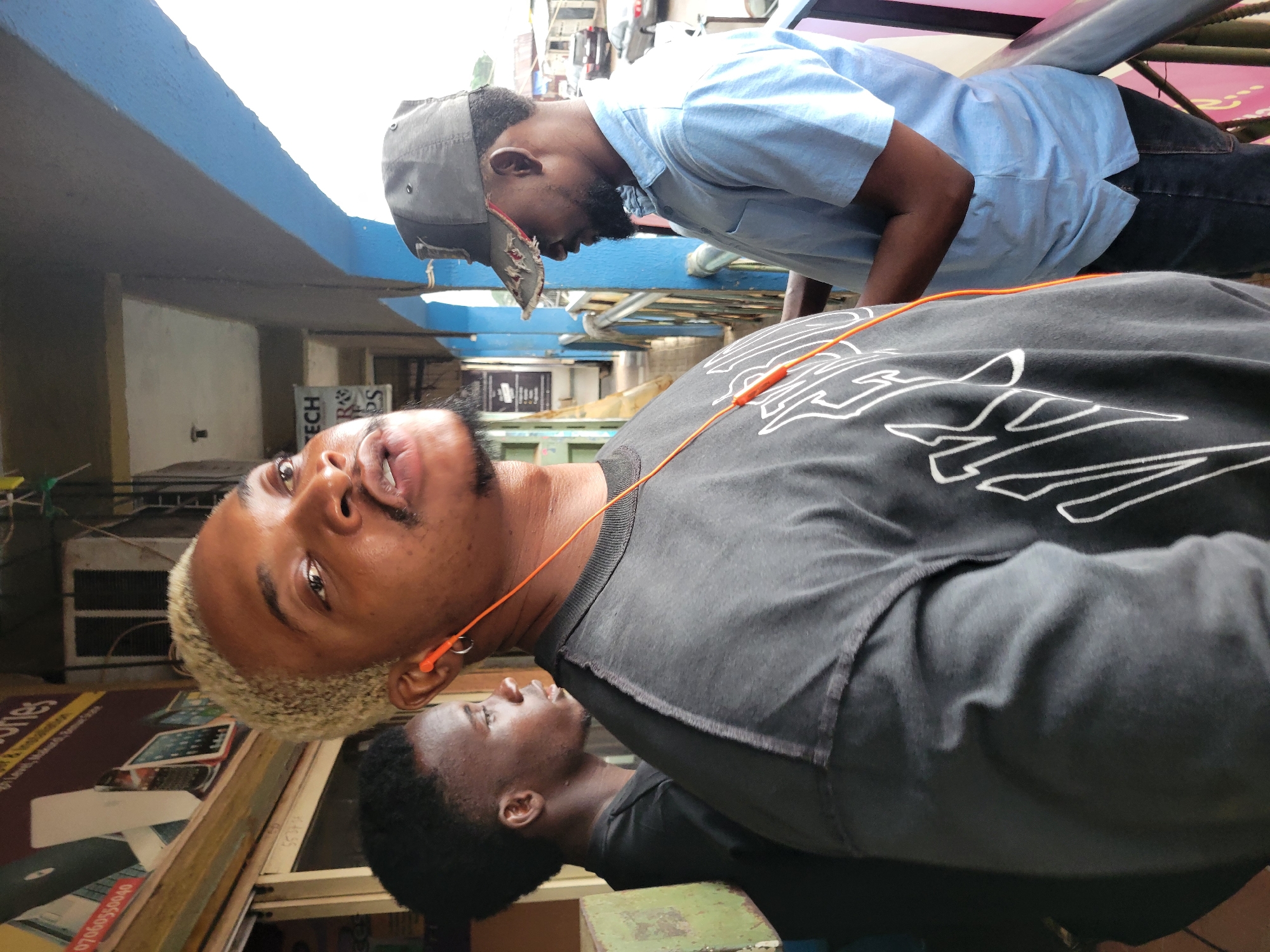 The photo captures three young men in what appears to be a commercial area of Makurdi, Nigeria. One man, facing the camera, has bleached blonde hair and is wearing a black shirt with white graphic designs and orange earphones. Another man stands behind him, wearing a black shirt. A third man, partially visible in the background, wears a light blue button-down shirt and a grey baseball cap. The setting includes the facade of shops and buildings, hinting at an urban commercial district. A sign for "TECH" can be seen in the background.