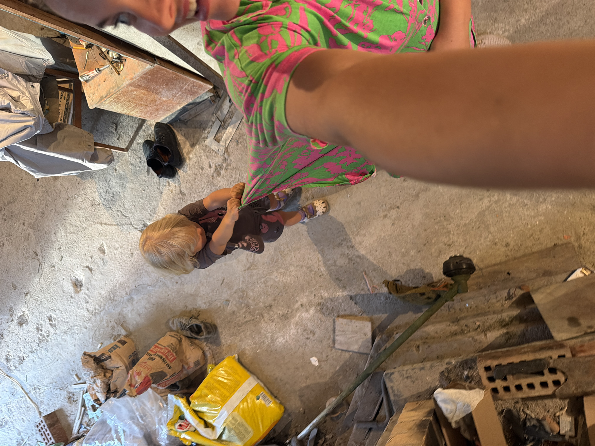An overhead, high-angle, medium shot captures an indoor scene with a concrete floor and dimly lit surroundings. A young child with blonde hair, wearing a dark t-shirt and shorts, stands in the center of the frame, holding onto a green and pink patterned fabric. The fabric appears to be part of a garment being pulled by the child. To the right of the child, an adult's arm and torso are visible, also in a green and pink patterned shirt, suggesting a selfie-style capture. The background features several objects, including bags of what appear to be construction materials, a pair of black shoes, a wooden chair with a gray jacket draped over it, a wooden box with tools on top, and parts of a weed trimmer. The overall impression is a casual, possibly domestic or workshop environment. The location in Baranovka, Russia is not visually indicated by the image content.