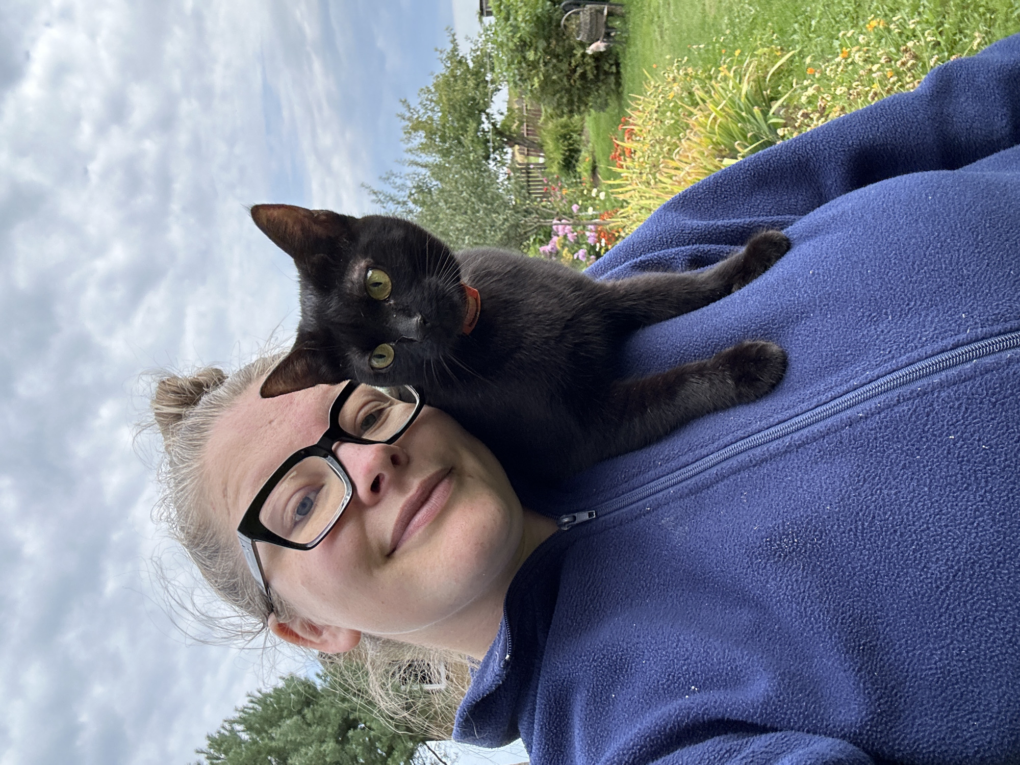 A woman with blonde hair in a bun and glasses is taking a selfie outdoors. A black cat with green eyes is perched on her left shoulder. The woman is wearing a blue fleece jacket with a zipper. The background shows a garden with flowers, green grass, and trees under a cloudy sky. The location is the village of Kalitsino, Russia.