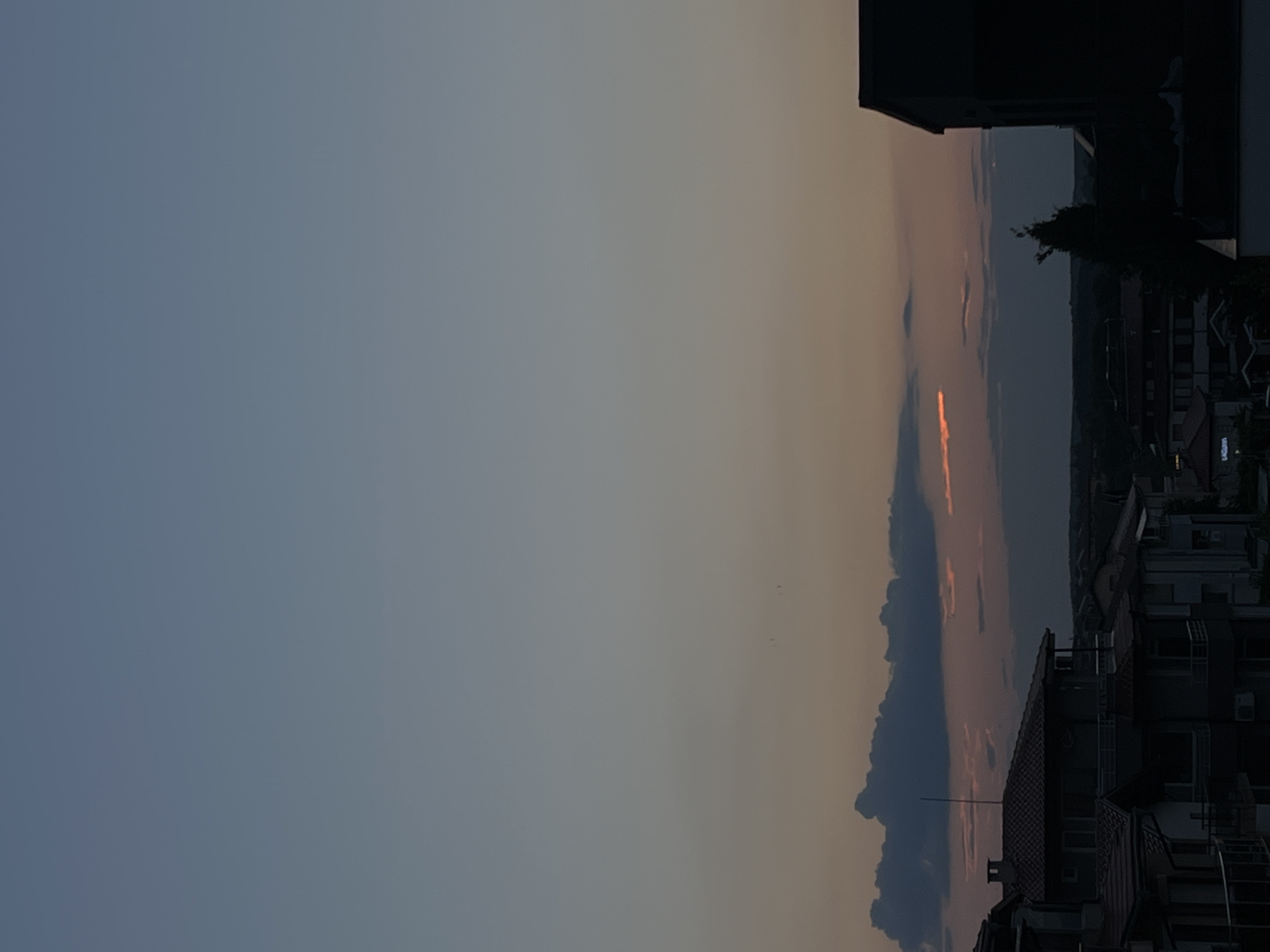 The image depicts a twilight scene over the city of Lozenets, Bulgaria. The sky transitions from a pale blue at the top to a soft orange and grey near the horizon, indicating the sun has recently set. Several dark, textured clouds are visible, some with a faint orange glow from the setting sun. Two small dark shapes, possibly birds, are in flight in the upper part of the sky.
In the foreground and mid-ground, the silhouettes of residential buildings are apparent. These structures appear to be multi-story, with visible rooftops and architectural details like balconies. One building on the right side of the image is a dark, imposing silhouette. The overall impression is of a quiet evening settlement. The environment is urban, characterized by the presence of housing and the fading light of day. There are no identifiable people or ongoing activities.