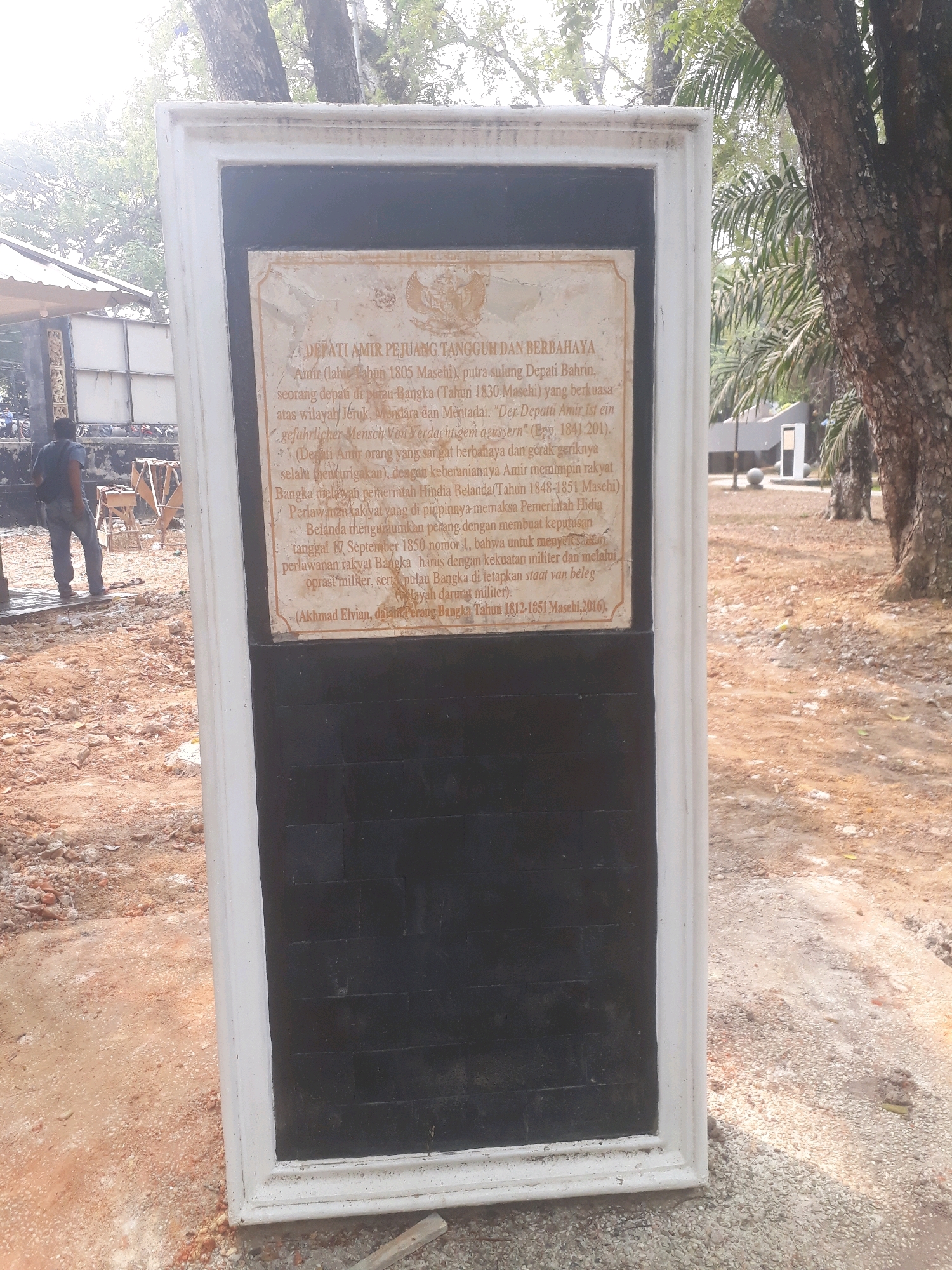 A weathered, rectangular stone plaque is mounted on a white stone monument, framed by trees and dry, dusty ground. The plaque, which is located in Pangkalpinang, Indonesia, features text and an emblem, possibly depicting a bird. The text appears to be historical, written in Indonesian and some German. The monument itself is placed on a patchy, gravelly surface with visible construction debris, suggesting an outdoor, possibly historical or memorial site. To the left of the monument, a dark-haired person wearing a dark shirt and grey trousers stands with their back to the camera, facing away from the monument and towards a sheltered area with what looks like construction materials. The overall environment is sunny, with shadows cast by trees, and the vegetation is tropical.
