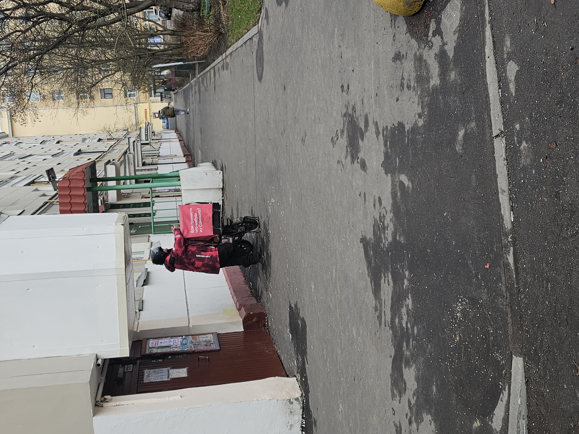 A delivery person wearing a red jacket, black pants, and a black balaclava stands next to a bicycle carrying a pink delivery bag near a building in Khimki, Russia. The building is a multi-story structure with white walls and a red and green awning. A sidewalk runs alongside the building. A second person walks farther down the sidewalk. Trees and other buildings are visible in the background.