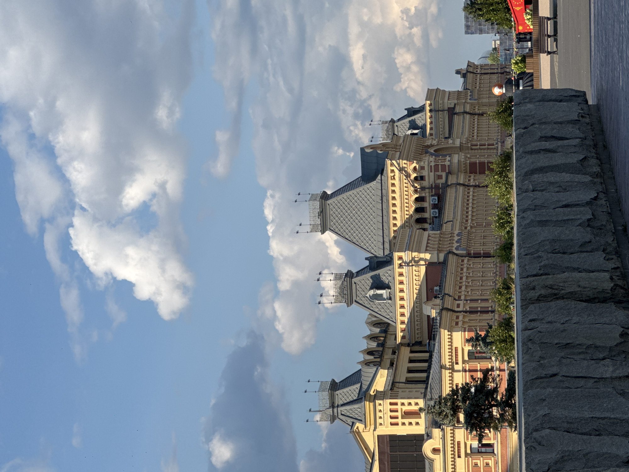 This is a daytime, eye-level shot of a grand, historic building in Nizhny Novgorod, Russia. The architecture is characterized by its intricate detailing, ornate facades in yellow and red, and a series of dark, pitched roofs topped with decorative finials and flags. The building appears to be multi-story with numerous windows and arched entrances. In the foreground, a rough-hewn stone wall or barrier runs horizontally across the frame, obscuring the lower portion of the building. Beyond the building to the right, there are glimpses of a public square with a wooden bench, some greenery, and what appears to be an art installation with a large, spherical, orange and white head. The sky above is a bright blue, dotted with scattered, fluffy white clouds. The lighting suggests it is either morning or late afternoon, casting subtle shadows. There are no visible people in the immediate vicinity of the building or the foreground wall.