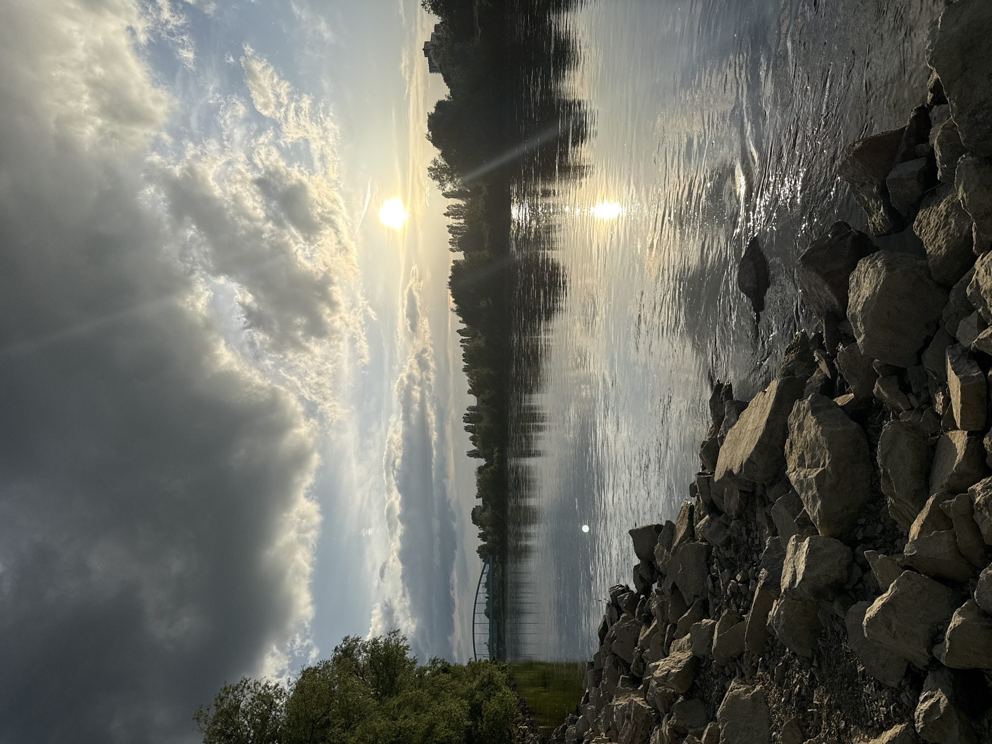A photograph captures a horizontal, wide-angle view of the Sava River in Zagreb, Croatia, under a dynamic sky. The sun, partially obscured by clouds, casts a bright reflection on the water’s surface. The river flows smoothly, with subtle ripples and variations in tone due to the sunlight. The riverbanks are densely lined with trees, appearing as dark silhouettes against the bright sky and water. A bridge is visible in the distance, spanning the river.
In the foreground, the right side of the frame is occupied by a rocky shoreline, composed of medium-sized, light-colored rocks. The left side features green foliage, likely trees or shrubs, adding a natural element to the scene. The sky is filled with clouds ranging from bright white to dark gray, creating a contrast.