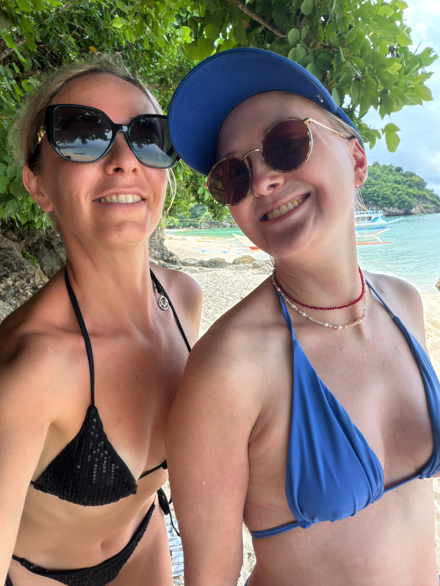 Two women wearing bikinis and sunglasses are taking a selfie on a beach. The woman on the left has blonde hair and is wearing a black sequined bikini. She is wearing large, black sunglasses. The woman on the right has blonde hair and is wearing a blue bikini and a blue baseball cap. She is wearing round sunglasses and a layered necklace. They are smiling at the camera. In the background, there is a sandy beach, green trees, and the ocean with boats. The location is identified as Malay, Philippines.