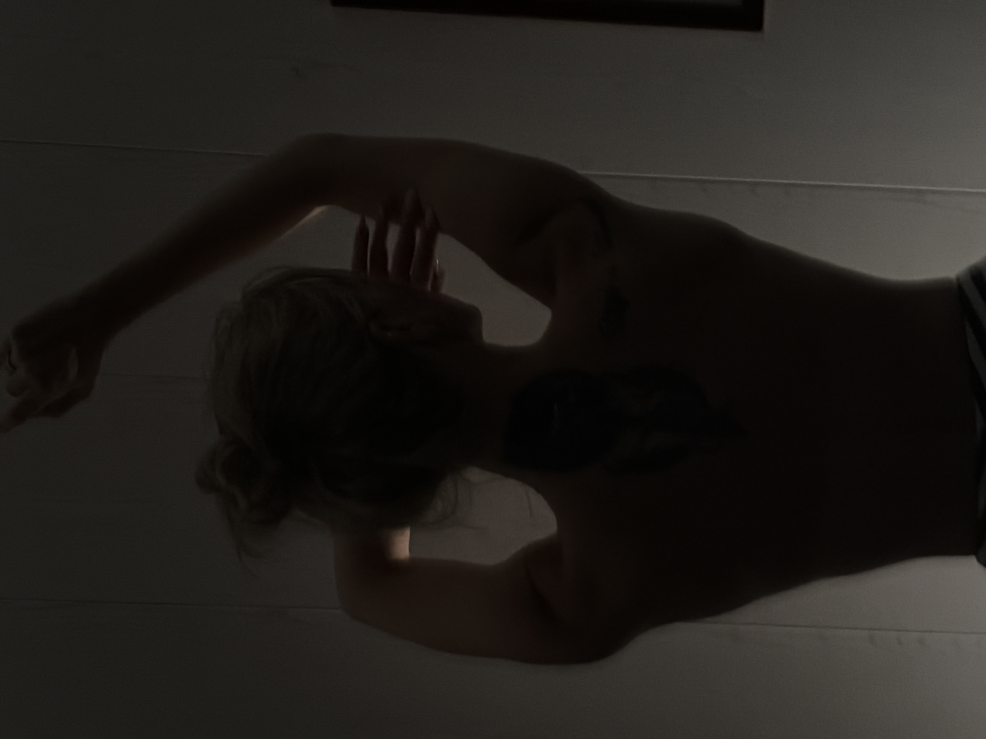 A person is lying on their side in a dimly lit interior. The person has blonde hair styled in a bun and a tattoo on their back. One arm is extended overhead, with the hand near their head. The other arm is out of view. The scene is likely indoors, given the wall and possibly a picture frame visible in the background. There is no confirmation this image was taken in Chaikovsky, Russia.