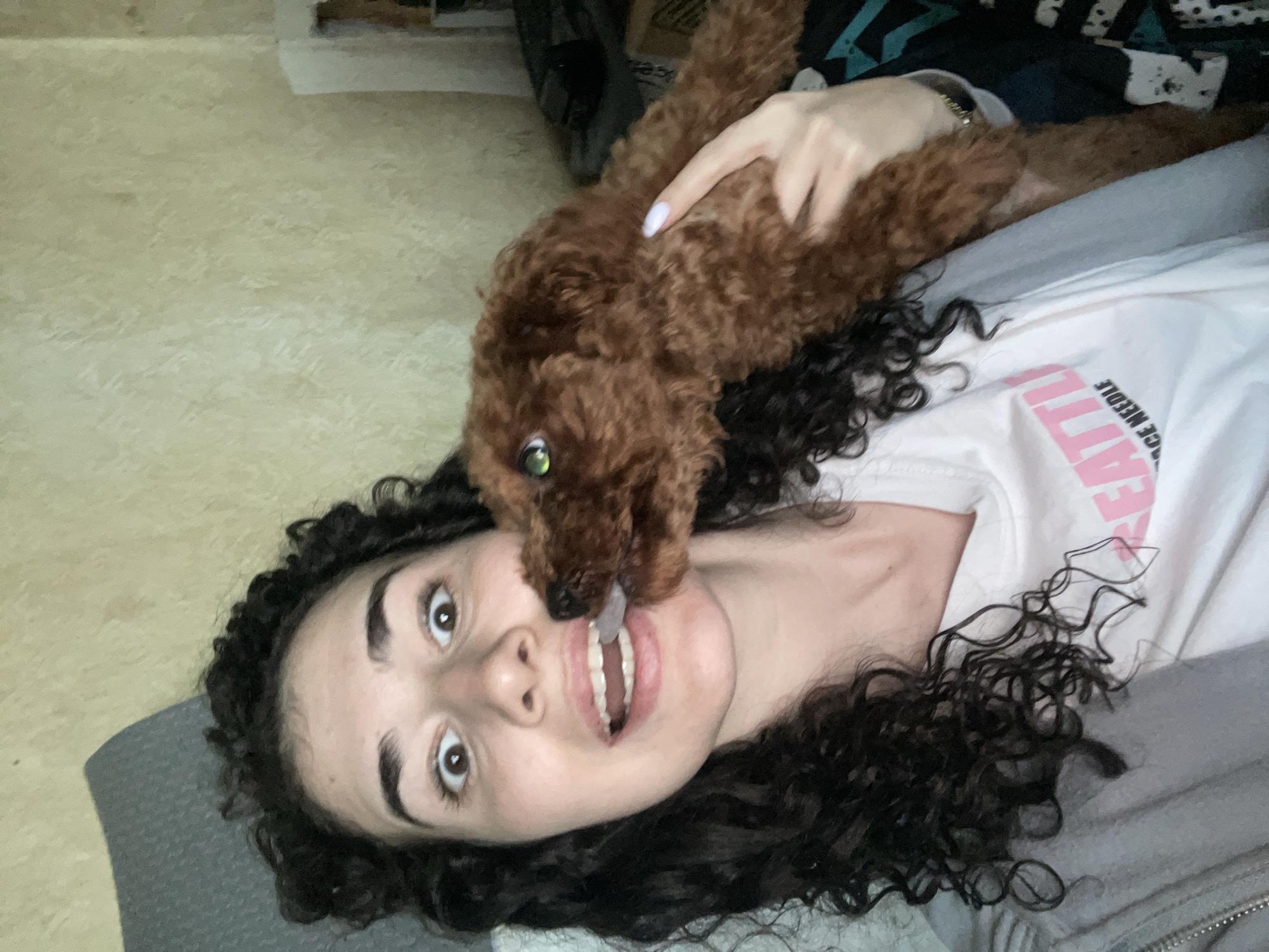 A woman lies on her back on a gray surface indoors. She has dark curly hair and is wearing a white T-shirt. A brown poodle is positioned on top of her, with its head near her face. The dog's mouth is near the woman's open mouth. A hand is on the dog's back. The floor has a light beige pattern. The city of Sofia, Bulgaria is not discernible in the image.