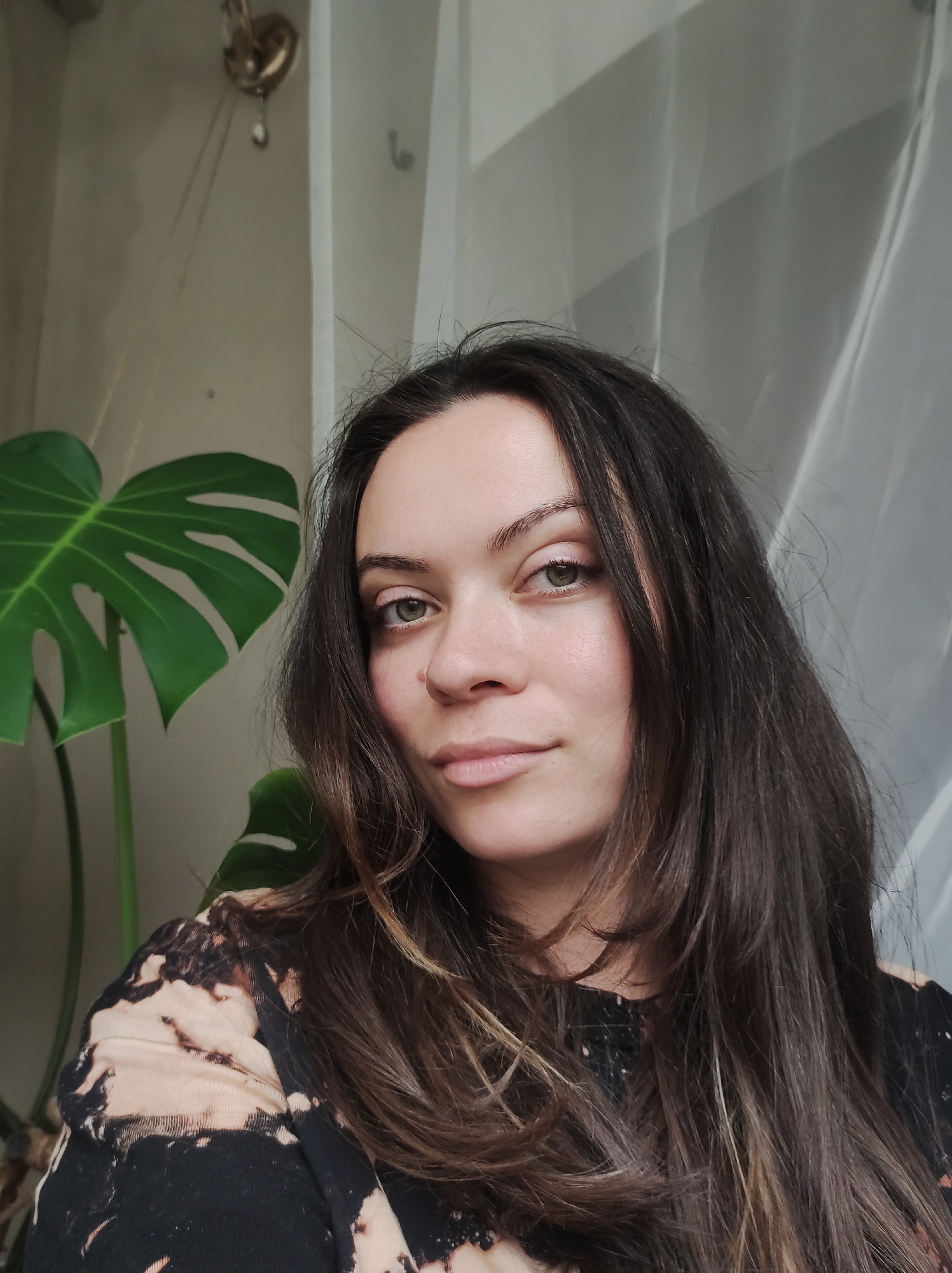 A medium, eye-level portrait shot of a woman in an indoor setting. The woman has dark brown hair with light brown highlights and fair skin. She has light green eyes and a nose ring in her left nostril. She is wearing a black and tan bleach-splattered long-sleeved shirt. To the left of the woman is a large Monstera plant with large, green leaves. Behind her is a white sheer curtain. In the upper-left corner, part of a gold-colored wall fixture can be seen.
The specific location of the photo within the city of Chaikovsky, Russia, cannot be determined.