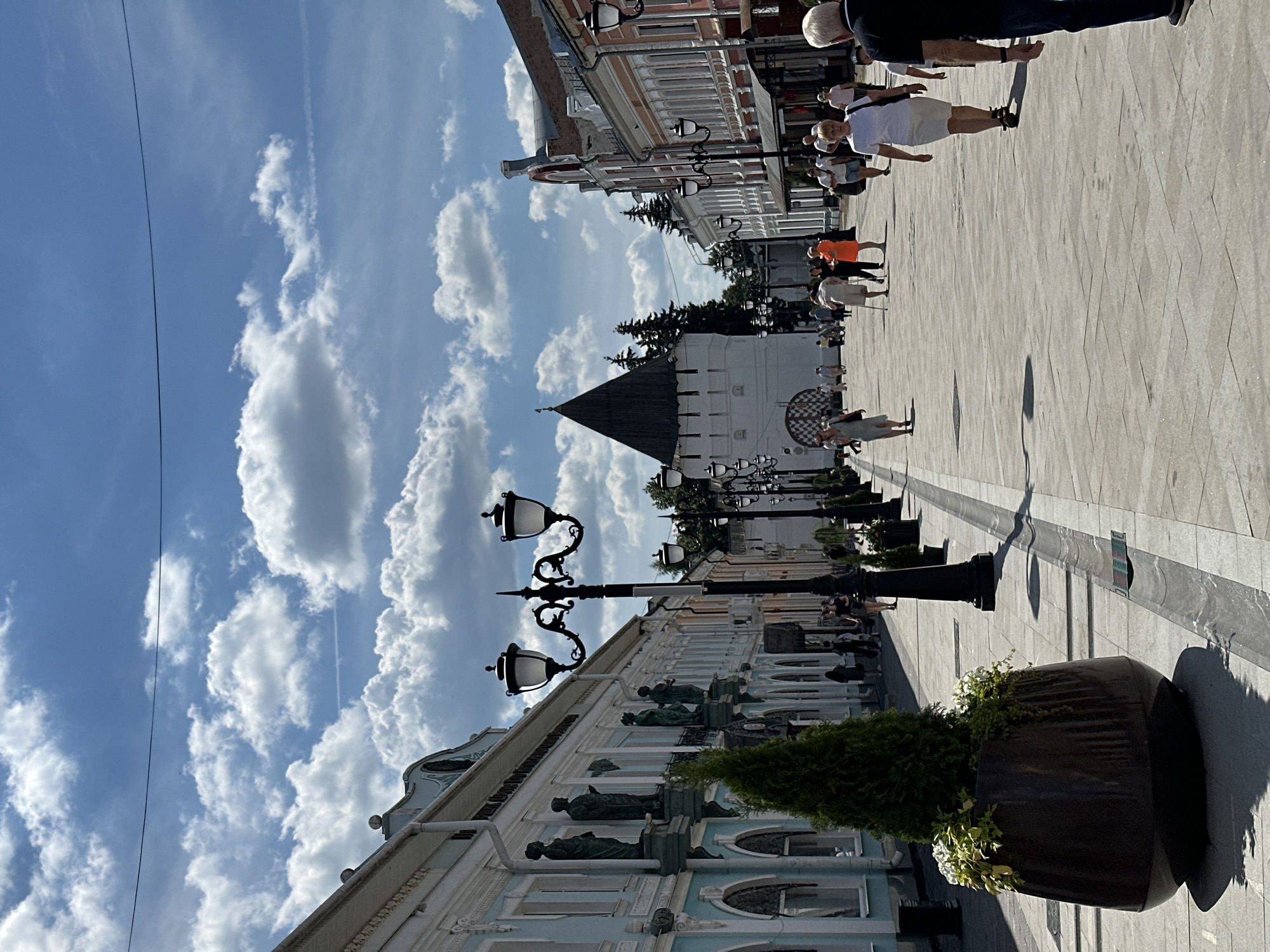 A street scene in Unknown, Unknown on a sunny day. A wide, paved pedestrian walkway is lined with buildings on either side. On the left, a light blue building with white decorative trim and statues on pedestals. A large, dark planter with a green conical shrub and white flowers sits in the foreground on the left. A black lamppost with two lights stands beside the planter. Across the street, buildings are painted in various colors, including peach and pink, with traditional architectural features. In the background, a white tower with a dark, conical roof, possibly part of a historical structure, is visible amongst trees. Several people are walking along the street. On the left, a woman in a light-colored dress walks away from the camera. In the center, a woman in an orange dress and an older woman with a cane are walking. Further down the street, more people are visible. A man in a black t-shirt and jeans is in the foreground on the right, partially obscuring the view. The sky is blue with scattered white clouds.